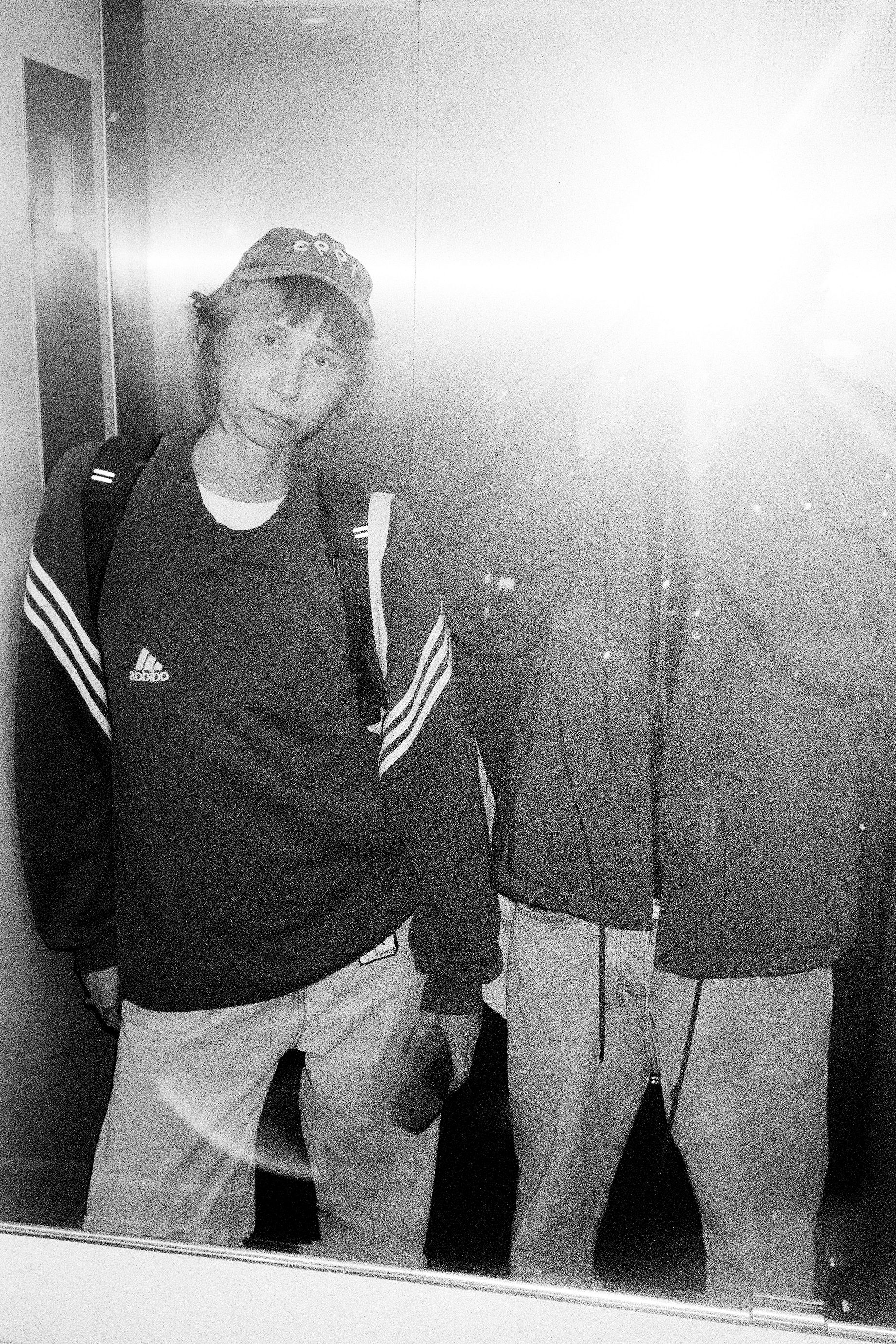 Czarno-białe zdjęcie zrobione w windzie przedstawia dwie osoby, z których jedna jest wyraźnie widoczna, ubrana w sportową bluzę i czapkę, a druga ma zasłoniętą twarz przez silne odbicie światła.
