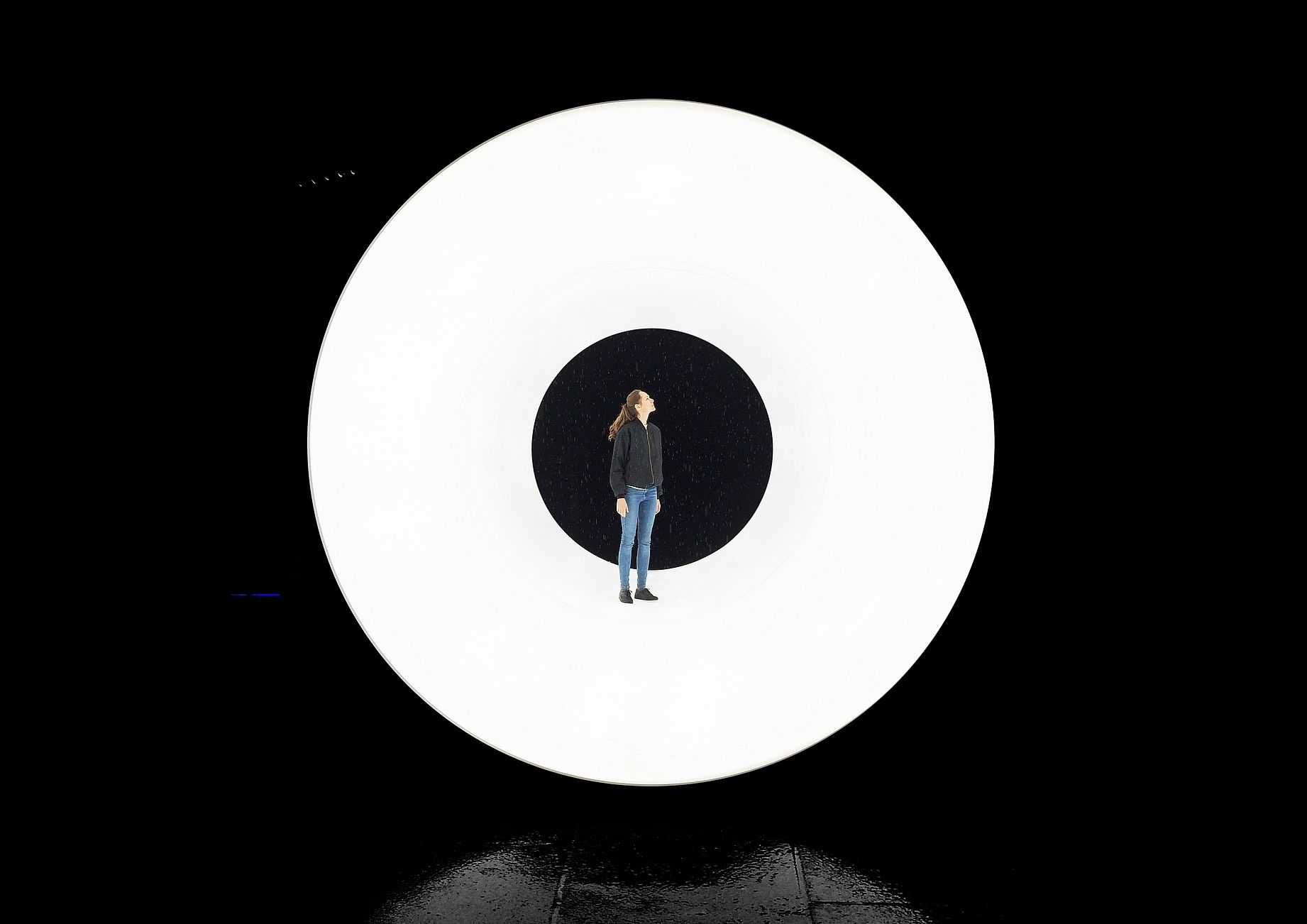 Na okrągłej, białej platformie, otoczonej całkowitą ciemnością, stoi postać zwrócona ku górze, wpatrując się w centralny, czarny otwór w środku koła.