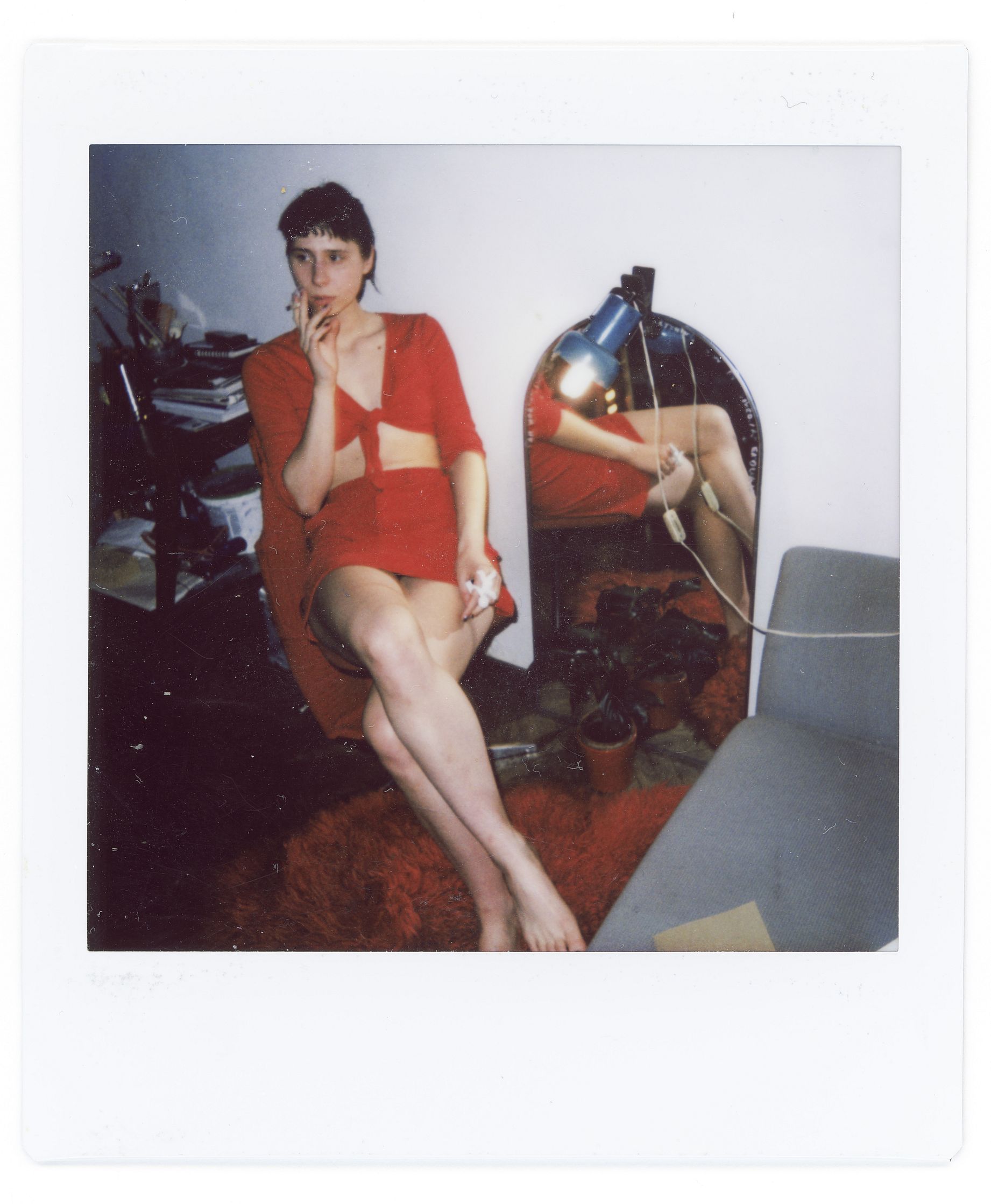 Zdjęcie przedstawia młodą kobietę w czerwonym, dwuczęściowym stroju, siedzącą boso na włochatym, czerwonym dywanie w pokoju z lustrem, w którym odbija się fragment jej nóg i lampa z niebieskim kloszem.