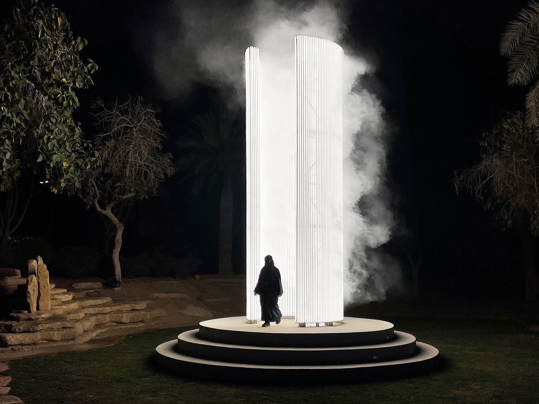 Na nocnym zdjęciu widać monumentalną, podświetlaną instalację świetlną Beacon Karoliny Hałatek, przypominającą cylindryczną latarnię morską, z której unosi się mgła, a przed którą na stopniach stoi postać w ciemnym ubraniu