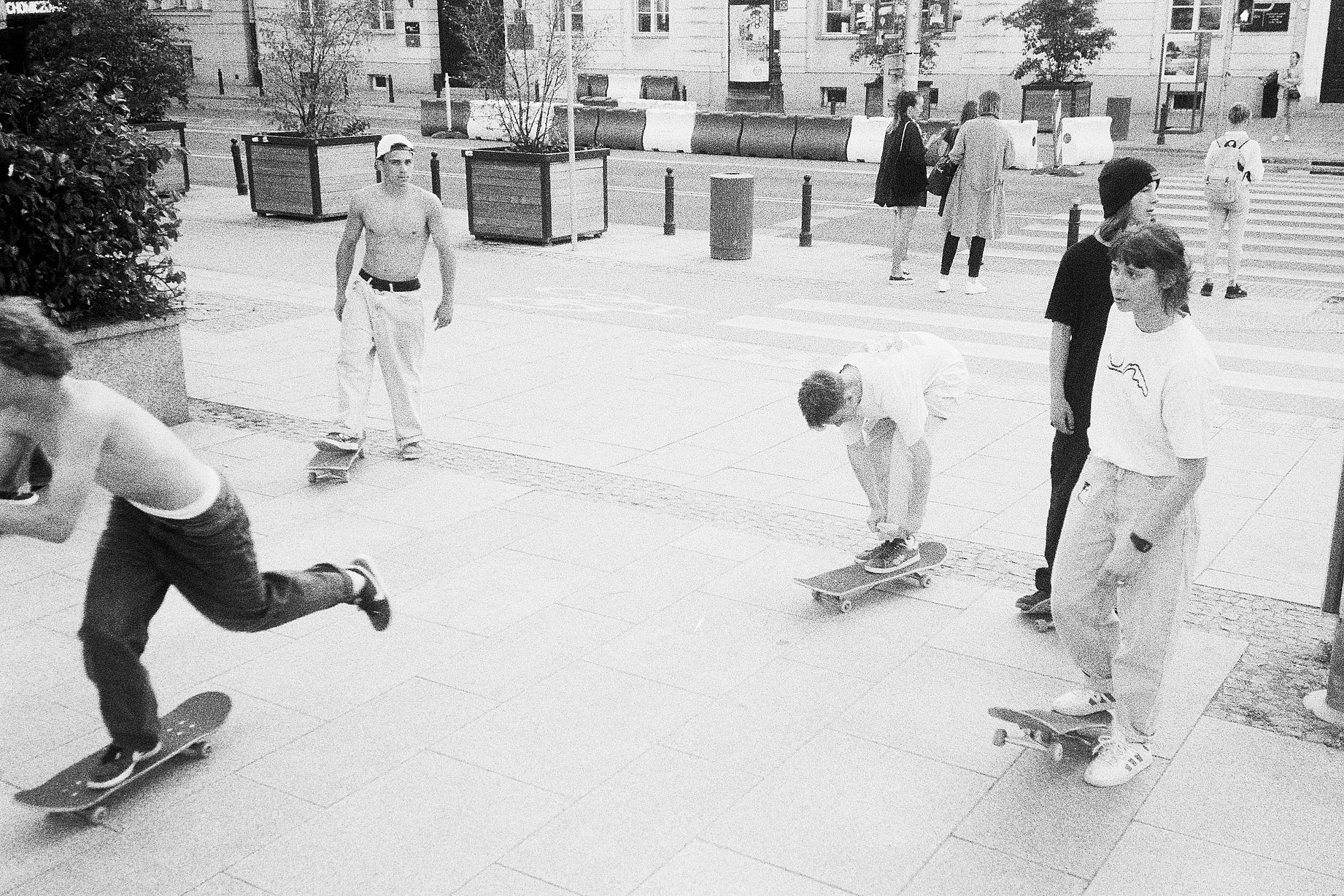 Czarno-białe zdjęcie plenerowe przedstawia grupę osób jeżdżących na deskorolkach na chodniku w miejskim otoczeniu, na którym widoczne są także inne postacie i elementy infrastruktury.