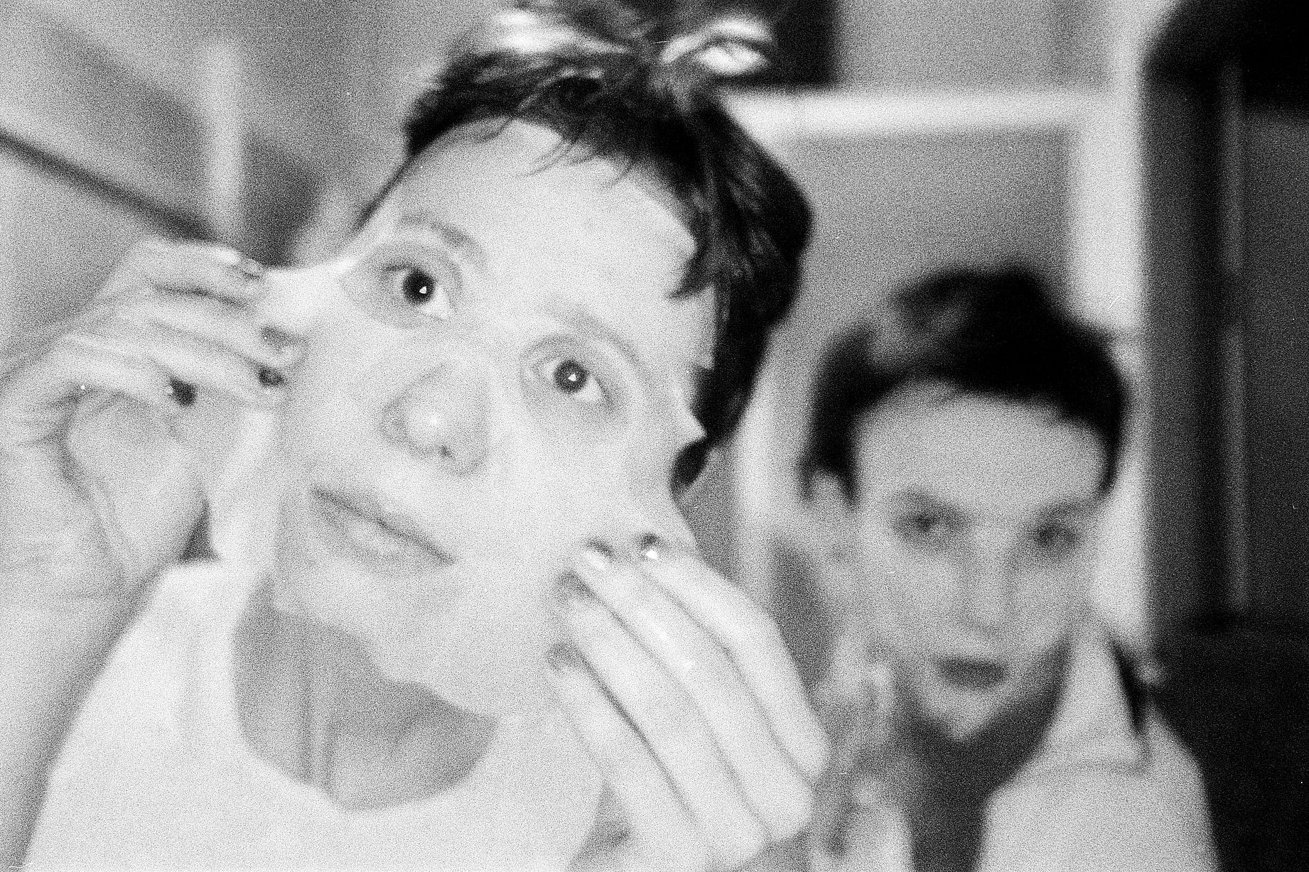 Czarno-białe zdjęcie przedstawia dwie osoby, z których jedna trzyma przy twarzy jasną maseczkę kosmetyczną, a druga jest z tyłu, nieostra.