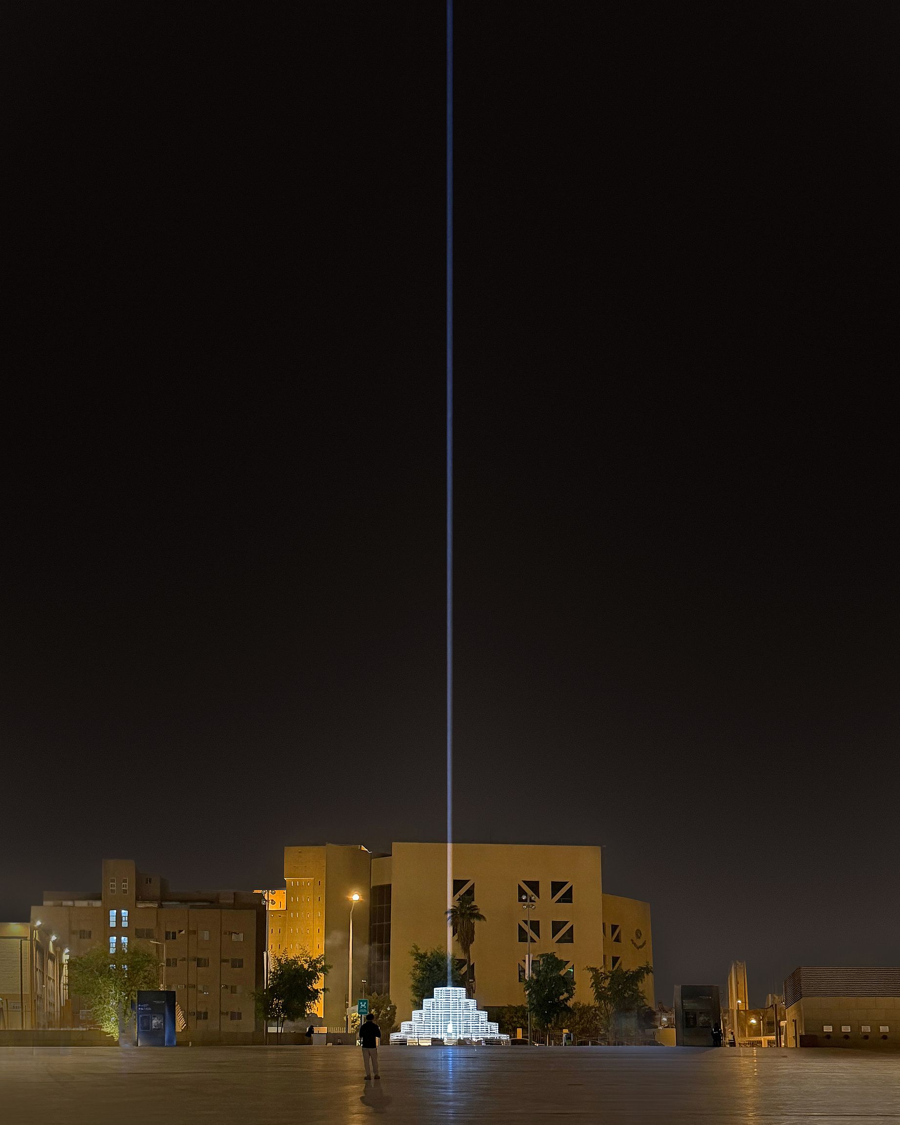 Piramidę z białych, podświetlanych skrzyń wieńczy pojedynczy, wąski promień niebieskawego światła, który o zmroku biegnie prosto w niebo, górując nad niższym, beżowym budynkiem o geometrycznych oknach, a na pierwszym planie znajduje się pusta, wybrukowana przestrzeń z małą postacią i kilkoma drzewami.