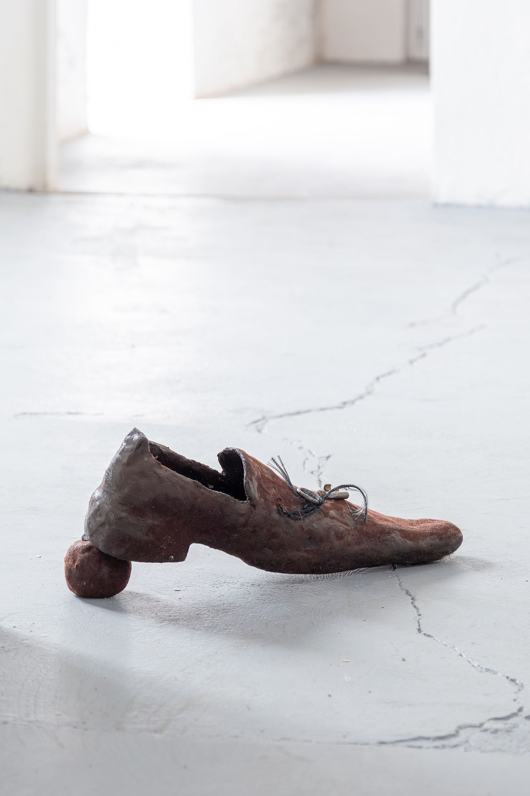Na zdjęciu widoczny jest pojedynczy, brązowy but o nietypowej, rzeźbiarskiej formie, z małym, okrągłym elementem przymocowanym do obcasa, leżący na betonowej podłodze.