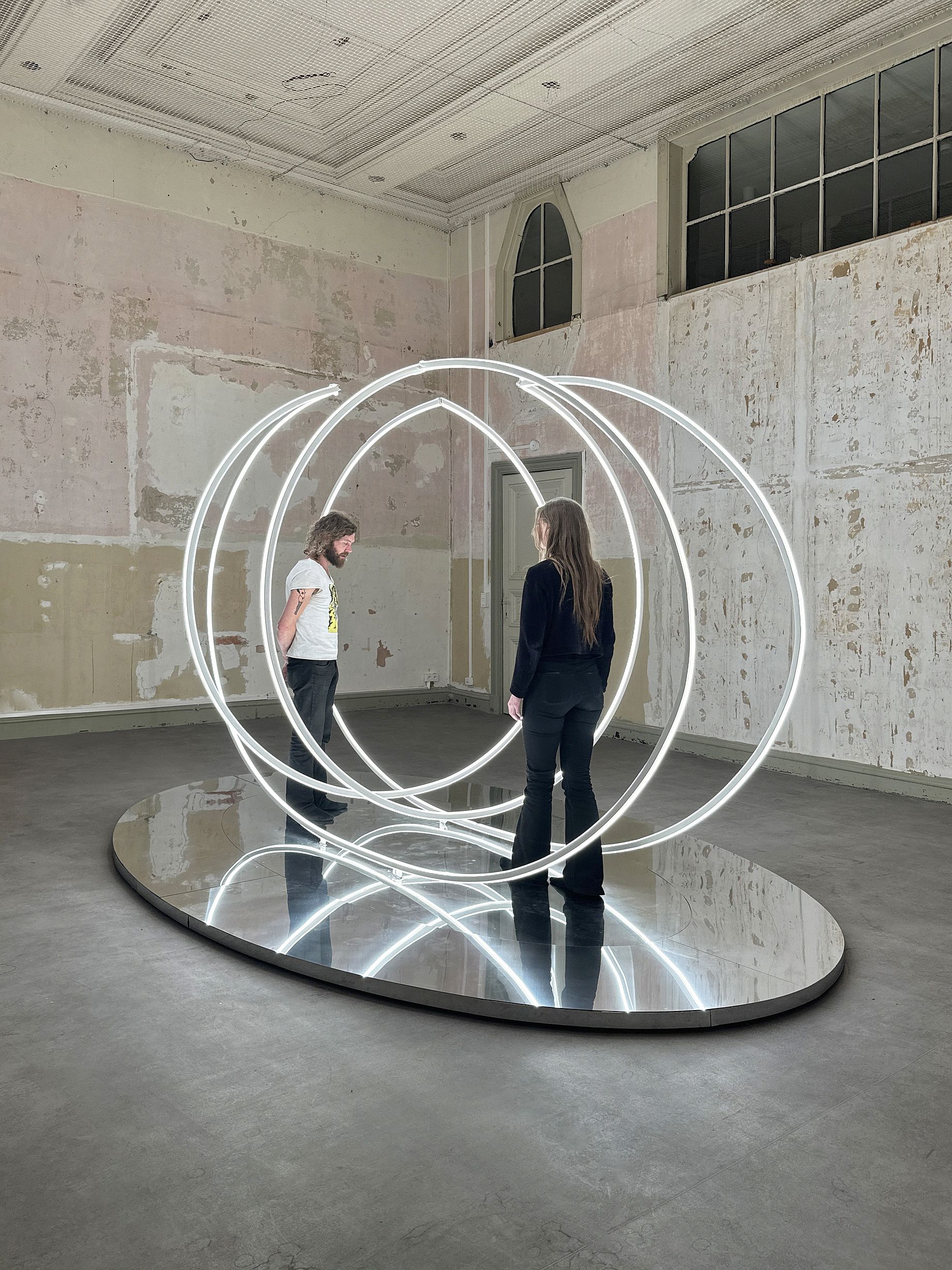 W pomieszczeniu o zniszczonych, łuszczących się ścianach i lustrzanej podłodze znajduje się instalacja artystyczna złożona z kilku nachodzących na siebie, białych, eliptycznych linii światła, wewnątrz których stoją dwie osoby.