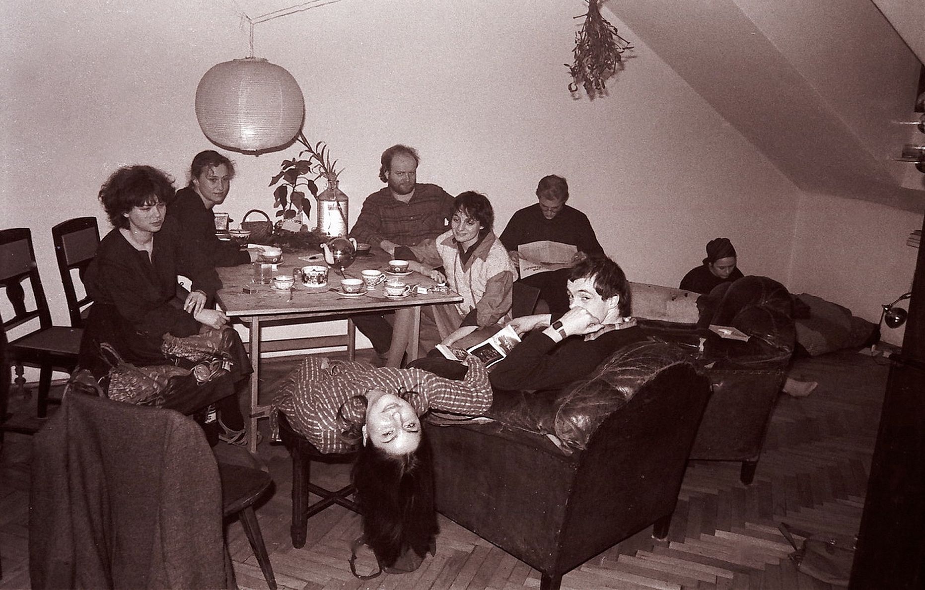Na czarno-białym zdjęciu widać grupę osób siedzących i leżących w niewielkim pokoju – część przy stole nakrytym filiżankami, część na kanapach, w swobodnej, towarzyskiej atmosferze.
