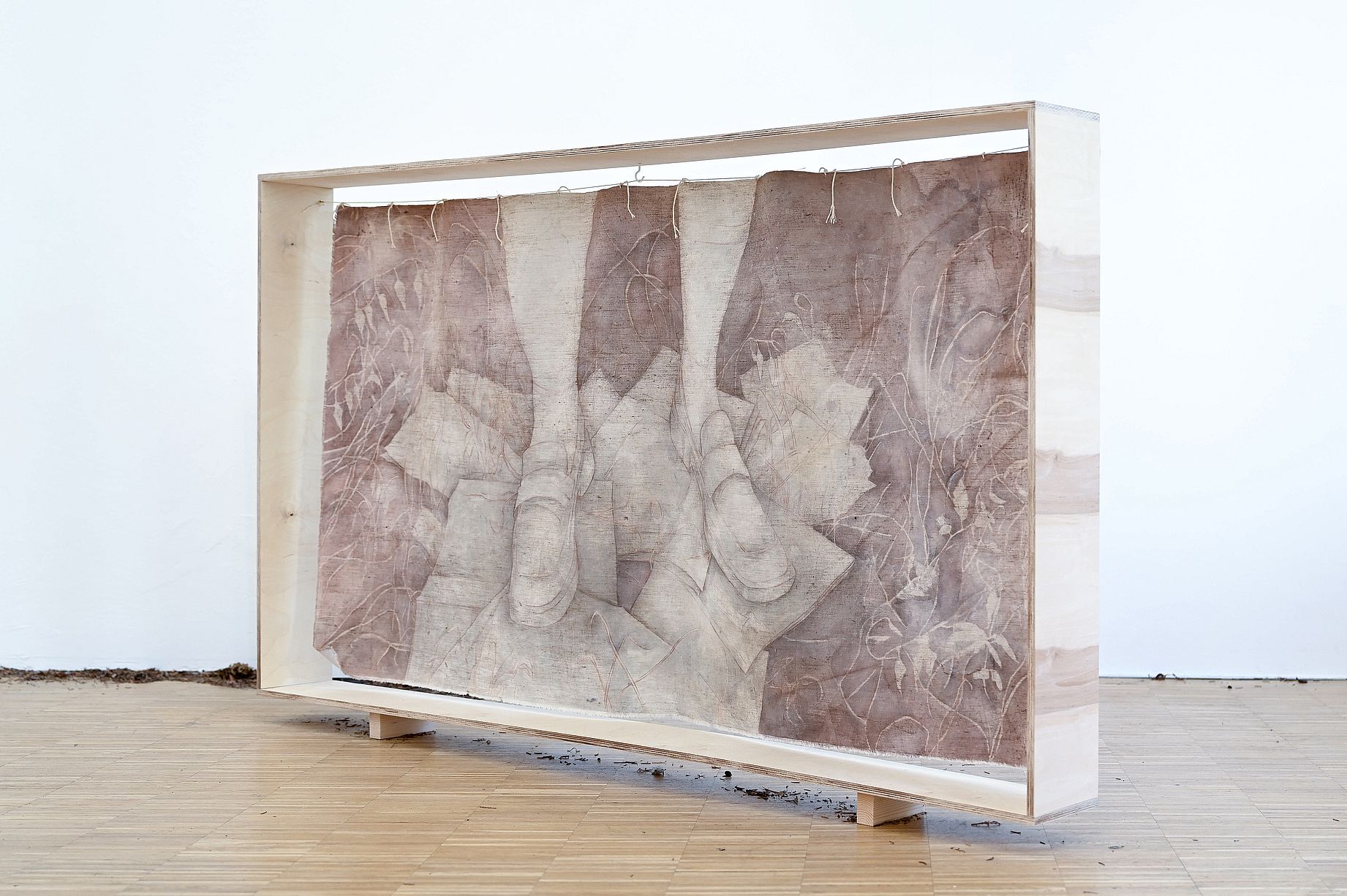 Prezentowana instalacja artystyczna, umieszczona w prostej, drewnianej ramie ekspozycyjnej, przedstawia wielowarstwową kompozycję z tkanin w odcieniach brązu i beżu, zawieszonych na sznurkach, z widocznymi abstrakcyjnymi wzorami przypominającymi korzenie lub mapy, leżącą na drewnianej podłodze z rozsypaną ziemią u podstawy.