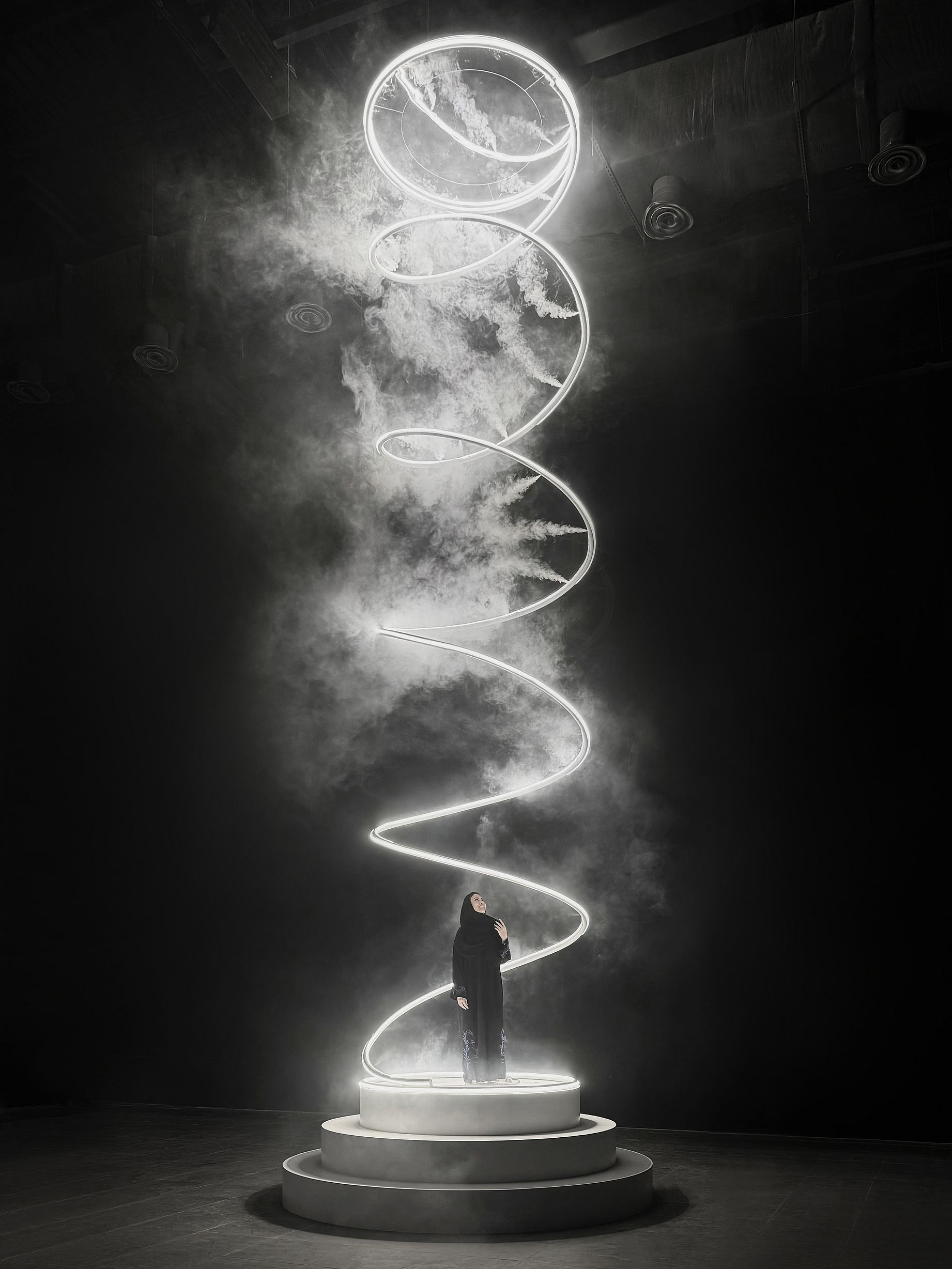Wysoka, świetlna instalacja w kształcie spirali, przypominająca trąbę powietrzną z mgły i białego światła LED, wznosi się z okrągłej podstawy, a pod nią, w centrum, stoi postać w ciemnym ubraniu.