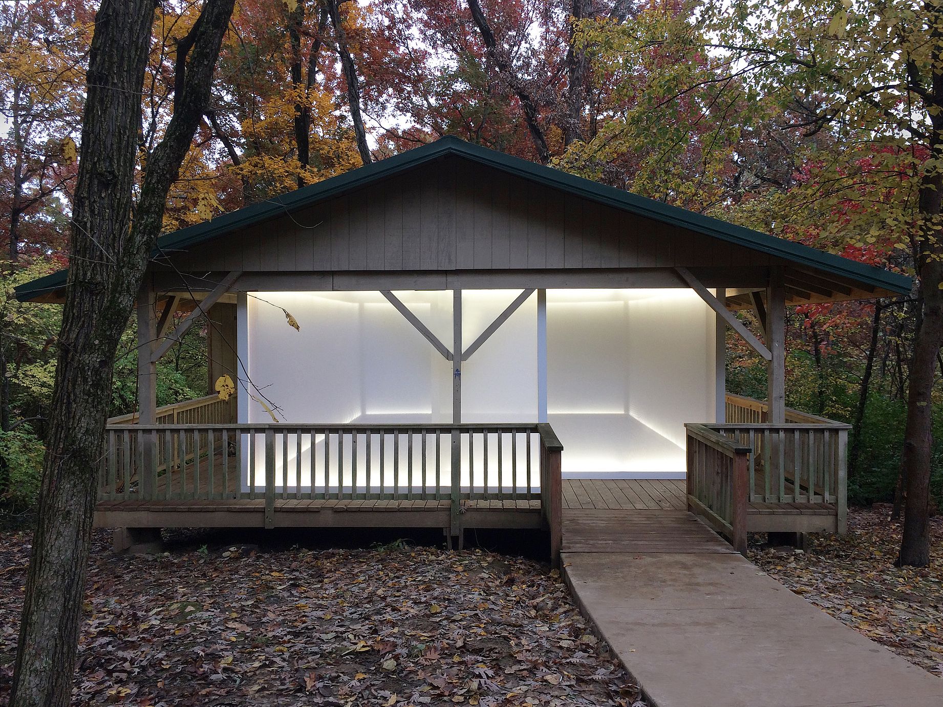  Mały, drewniany pawilon o otwartym boku, położony w jesiennym lesie wśród opadłych liści, ma wnętrze podświetlone jasnym, białym światłem zza białych, półprzezroczystych ścian, tworząc kontrast z otoczeniem, a na pierwszy plan wysuwa się drewniany podest z balustradą, do którego prowadzi betonowa ścieżka.