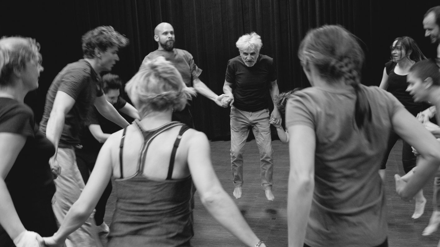 Grupa ludzi w różnym wieku trzyma się za ręce, tworząc krąg i wspólnie skacząc podczas energetycznego, radosnego spotkania w sali z ciemnymi zasłonami.