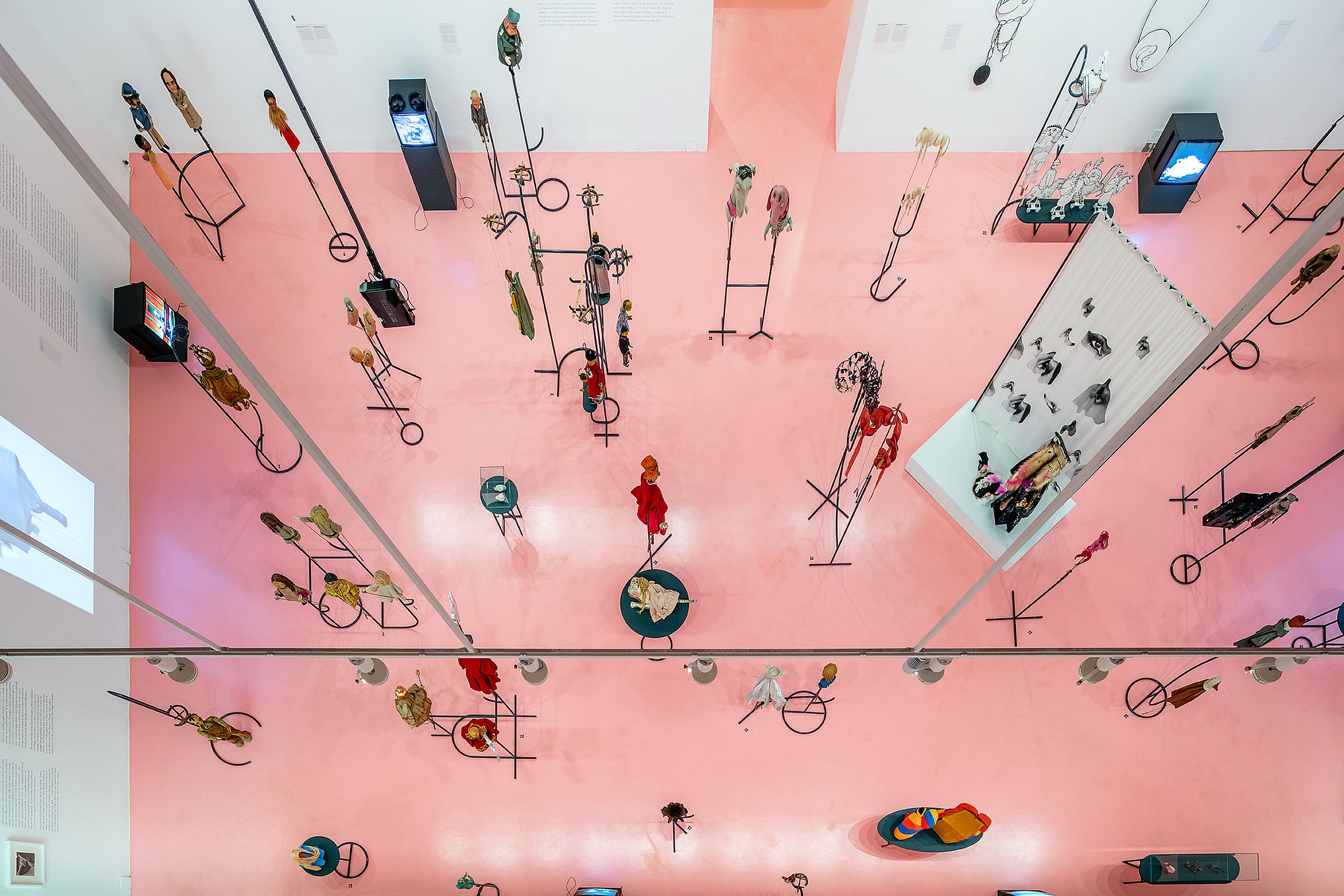 Zdjęcie przedstawia wystawę instalacji artystycznych, prawdopodobnie związanych z teatrem lalek lub sztuką kinetyczną, oglądaną z góry.  Wystawa ma miejsce w dużej, otwartej przestrzeni z podłogą w kolorze różowym. Prezentowane obiekty to małe figurki lub lalki zamontowane na metalowych stojakach lub prętach, rozmieszczone w różnych konfiguracjach.