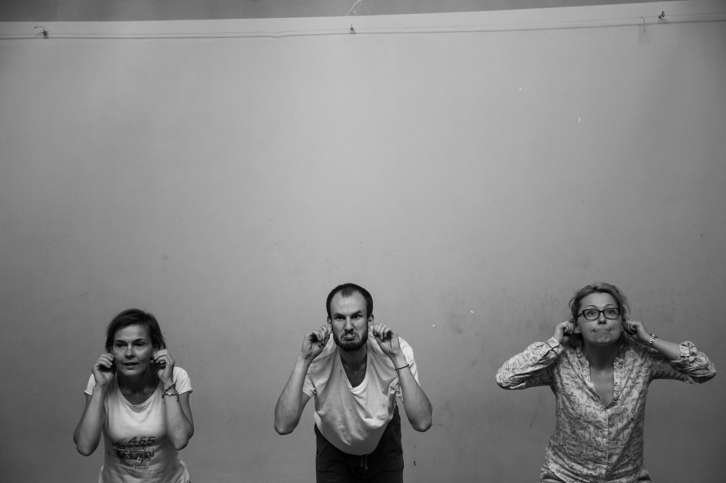 Na zdjęciu widać trzy osoby wykonujące zabawną minę i gest polegający na ciągnięciu się za uszy, jakby brały udział w ćwiczeniu teatralnym lub improwizacyjnym – stoją w jednej linii na tle pustej ściany, w skupieniu i z zaangażowaniem.