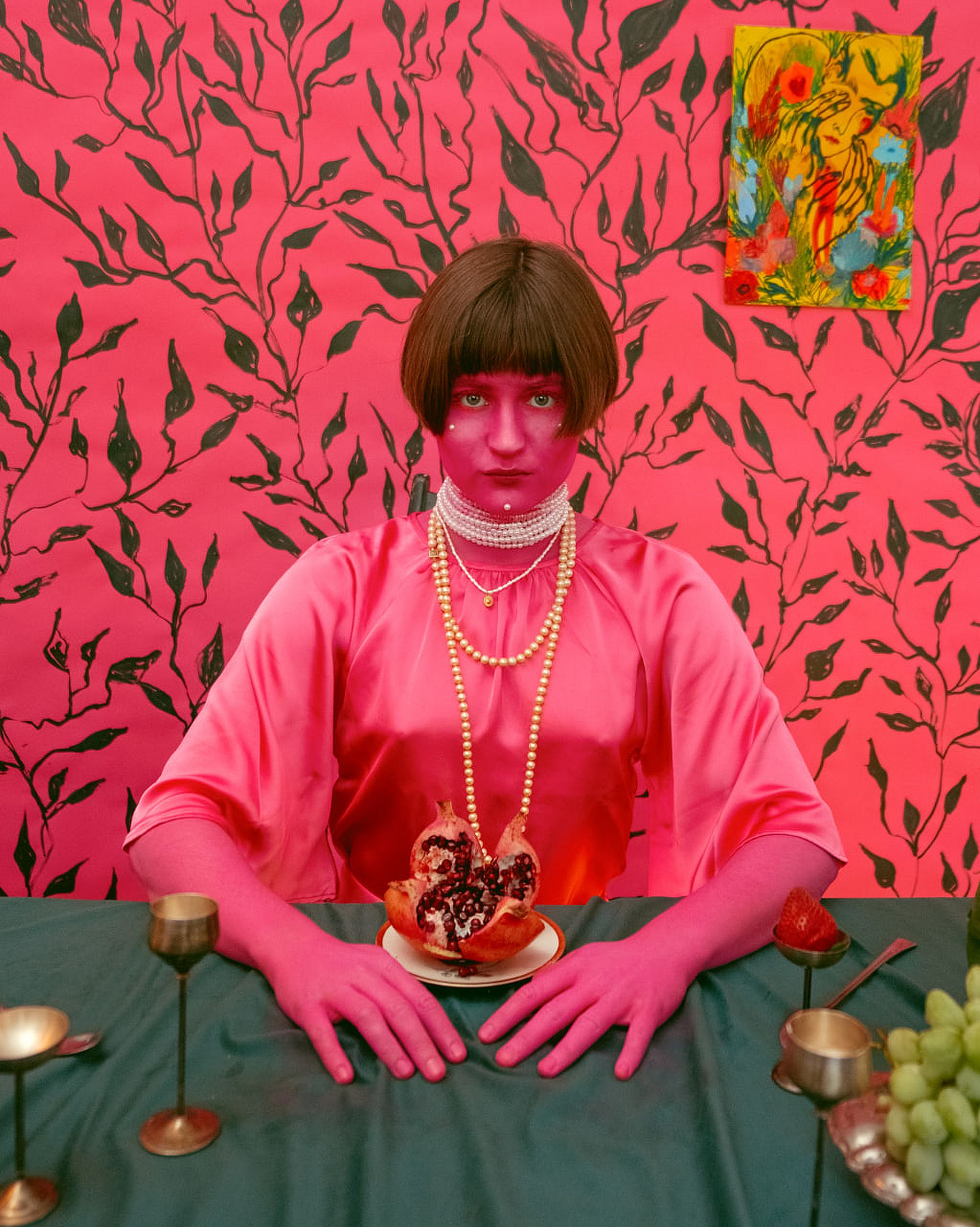 Kobieta ubrana na różowo, z pomalowaną czerwono twarzą siedzi przy stole, na którym stoją kielichy i owoce, w tle tkanina z roślinnym motywem