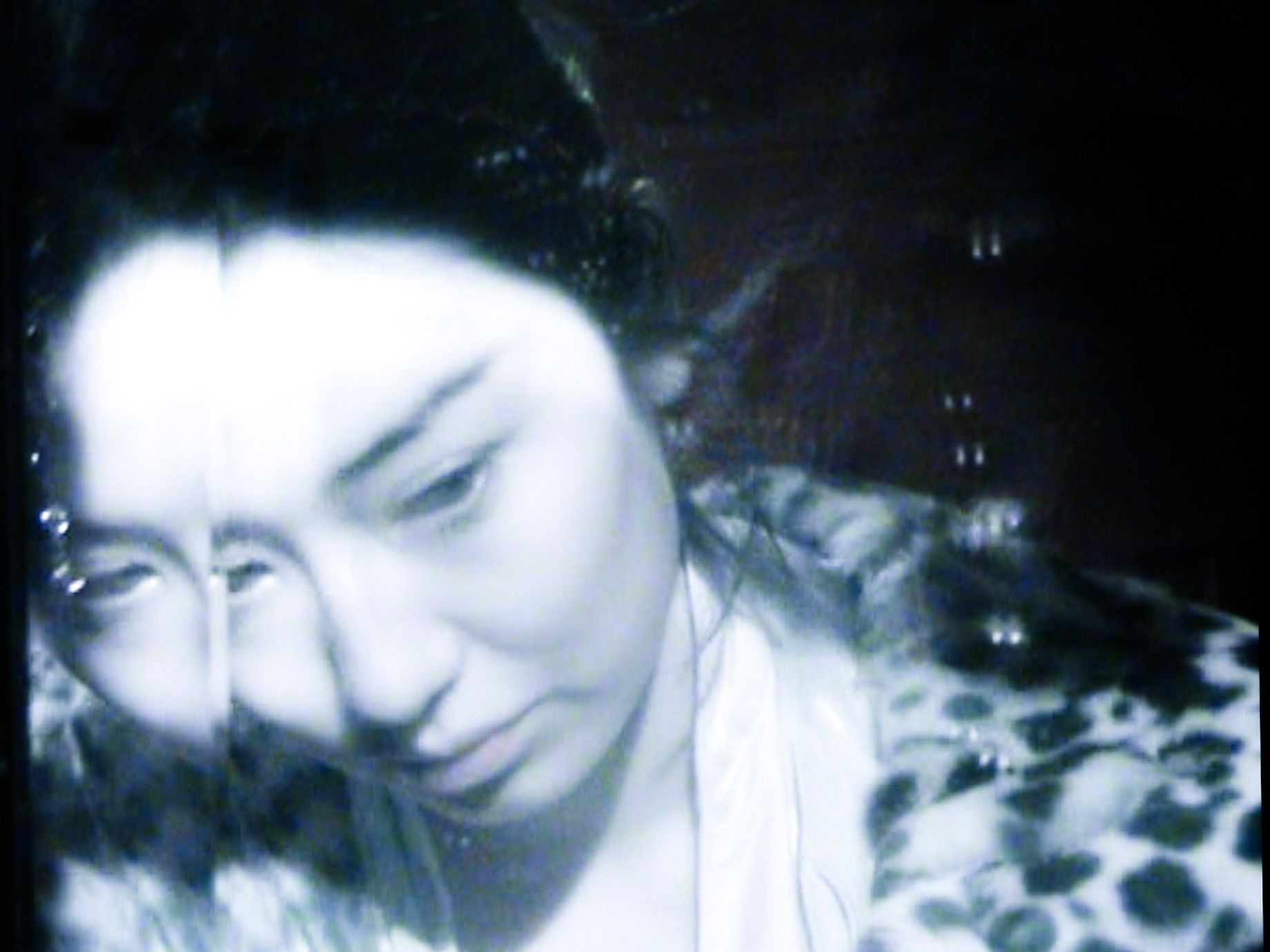 Zdjęcie przedstawia czarno-białe, ziarniste ujęcie twarzy młodej kobiety, która patrzy w dół, z częścią twarzy prześwietloną jasnym światłem, a resztą w cieniu, ubrana w odzież we wzór przypominający lamparta