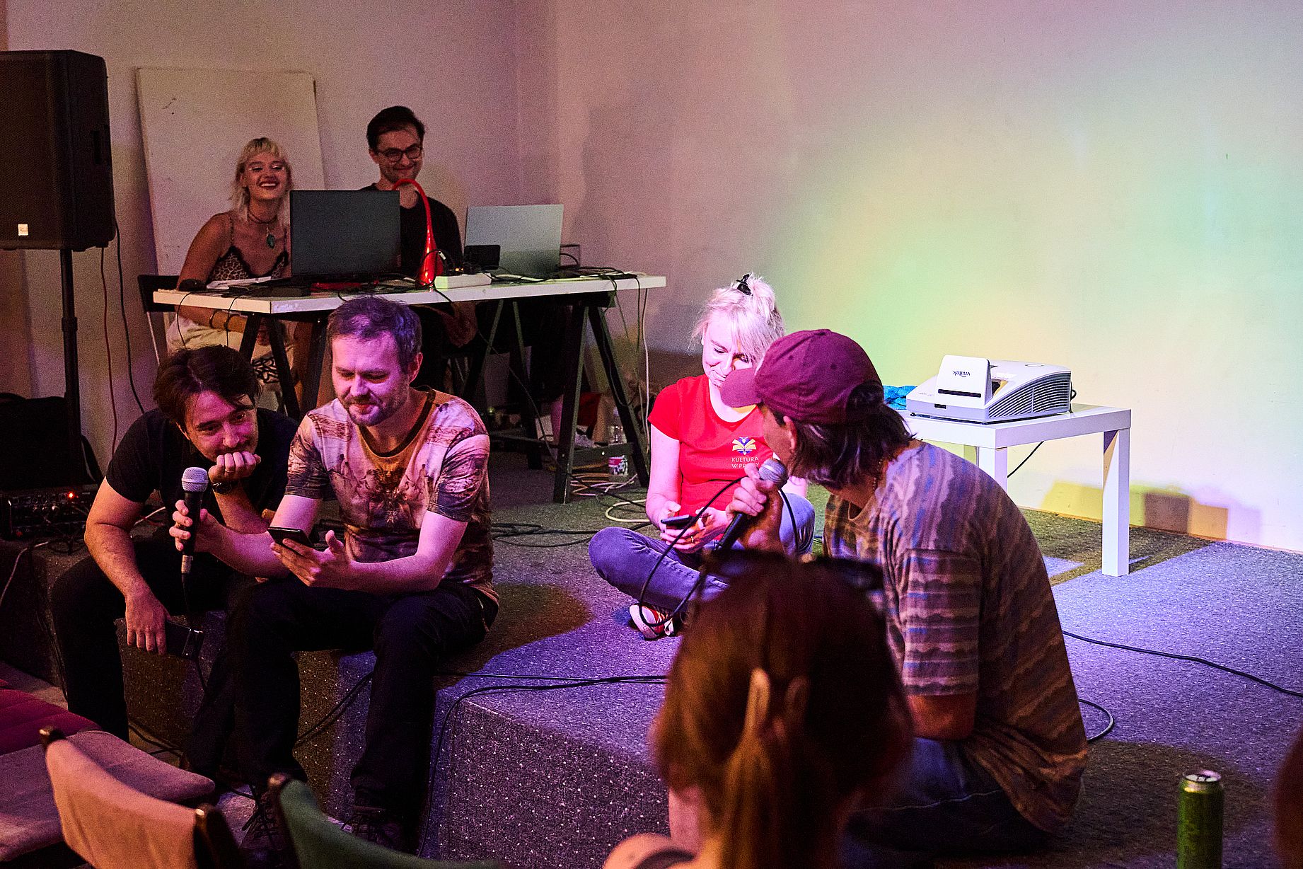 Na małej scenie siedzi kilka osób, dwi z mikrofonem. W tle dwie osoby przy stole z laptopami. To pewno jakieś turniej, spotkanie