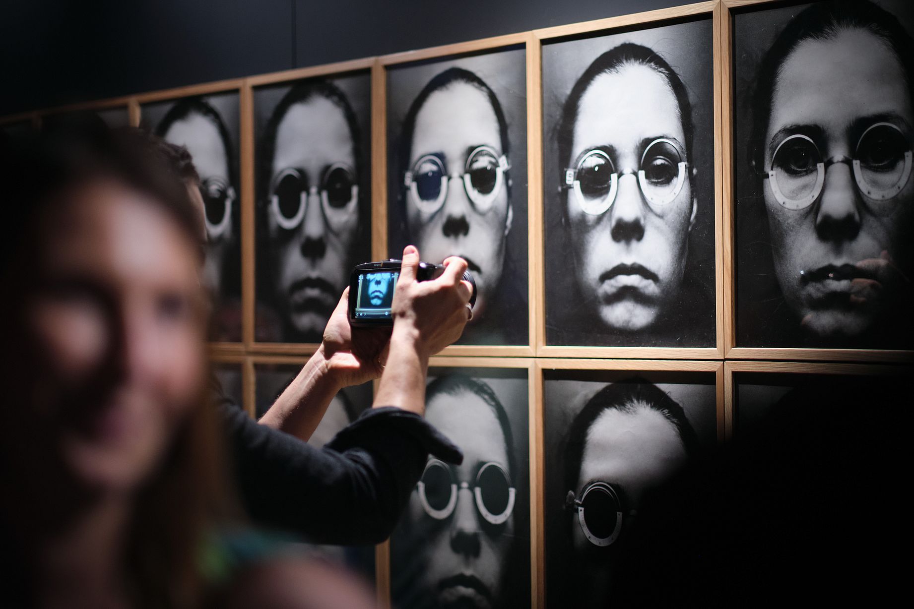 Widok wystawy, prawie to samo zdjęcie przedstawiające kobietę w okularach, w kilkunastu wersjach, ktoś ogląda zdjęcie w paracie fotograficznyme
