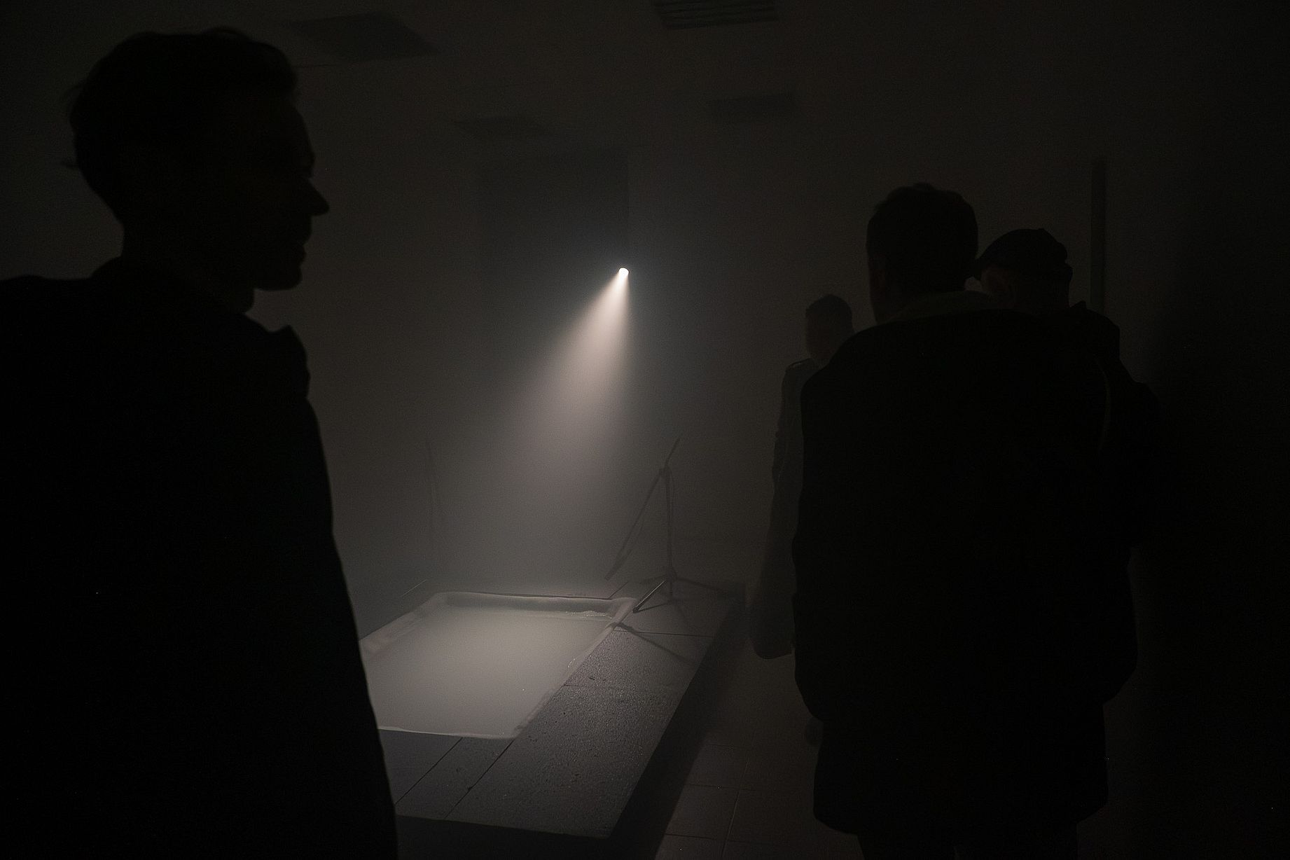 Zdjęcie przedstawia ludzi stojących w słabo oświetlonym pomieszczeniu, oświetlonym pojedynczym, jasnym snopem światła.