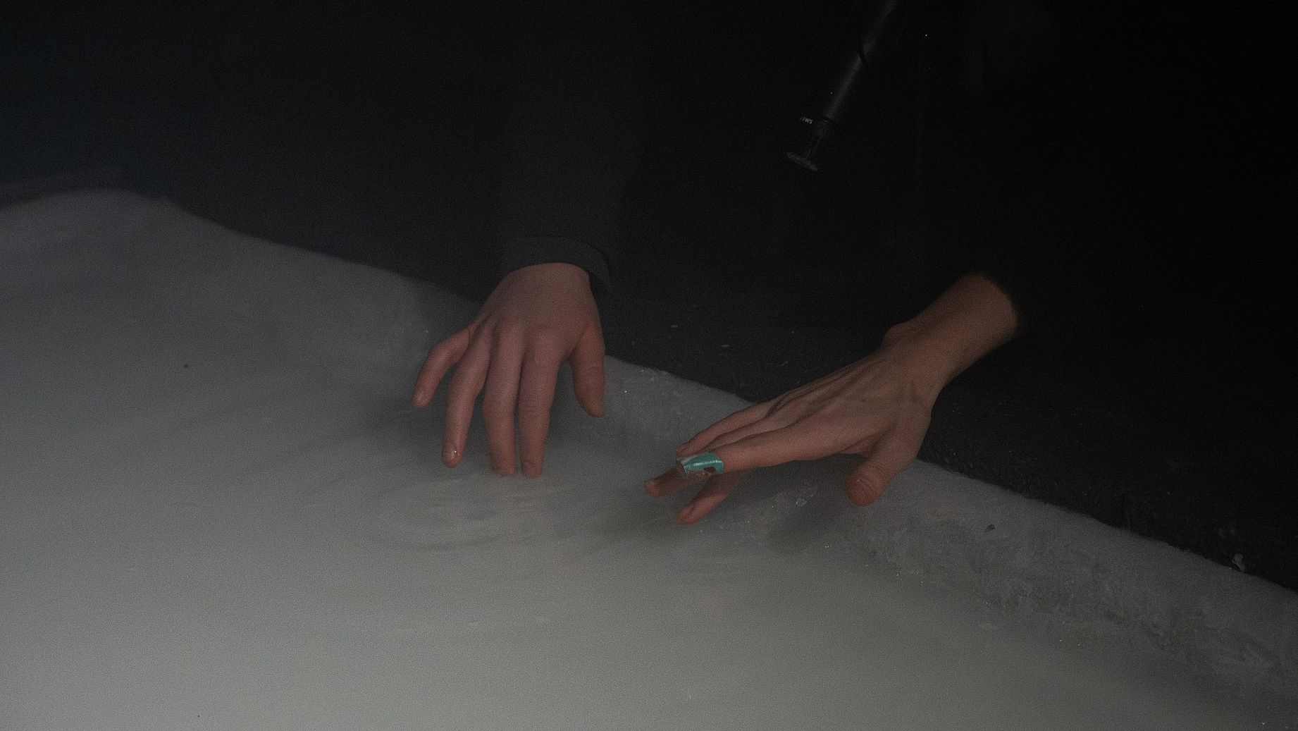 Zdjęcie przedstawia zbliżenie na dwie ludzkie ręce, prawdopodobnie kobiece, zanurzone w nieprzezroczystym, białym lub jasnoszarym płynie, który może być pianą lub mlekiem.