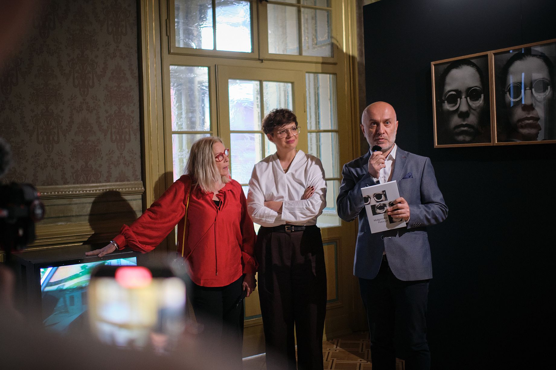 Otwarcie wystawy, kobieta w czerwonej bluzce, obok kobieta w białej  i mężczyzna w szarej marynarce