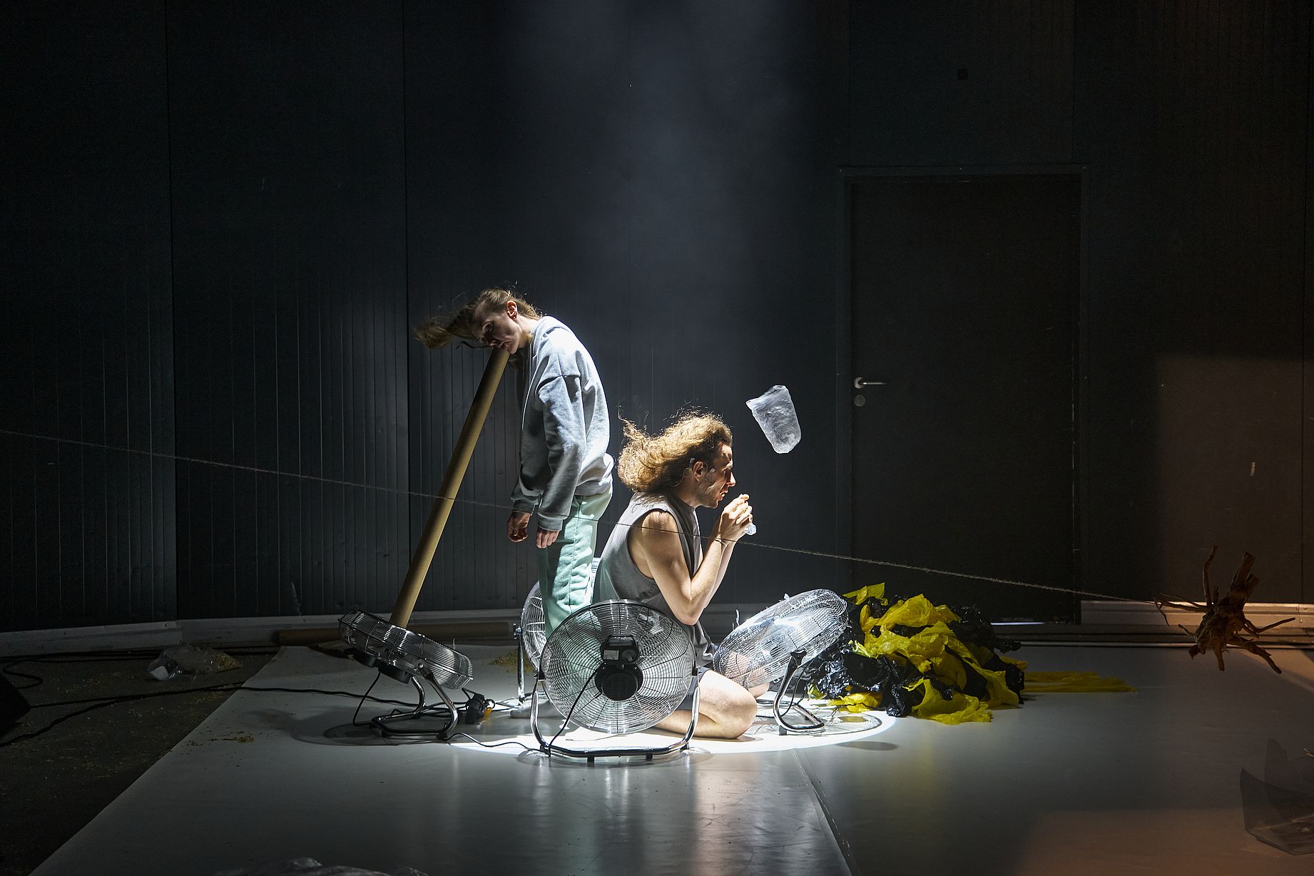 Zdjęcie przedstawia parę performerów na minimalistycznej, ciemnej scenie teatralnej, którzy klęczą i stoją w snopie jasnego światła pośród włączonych wentylatorów podłogowych mierzwiących ich włosy.