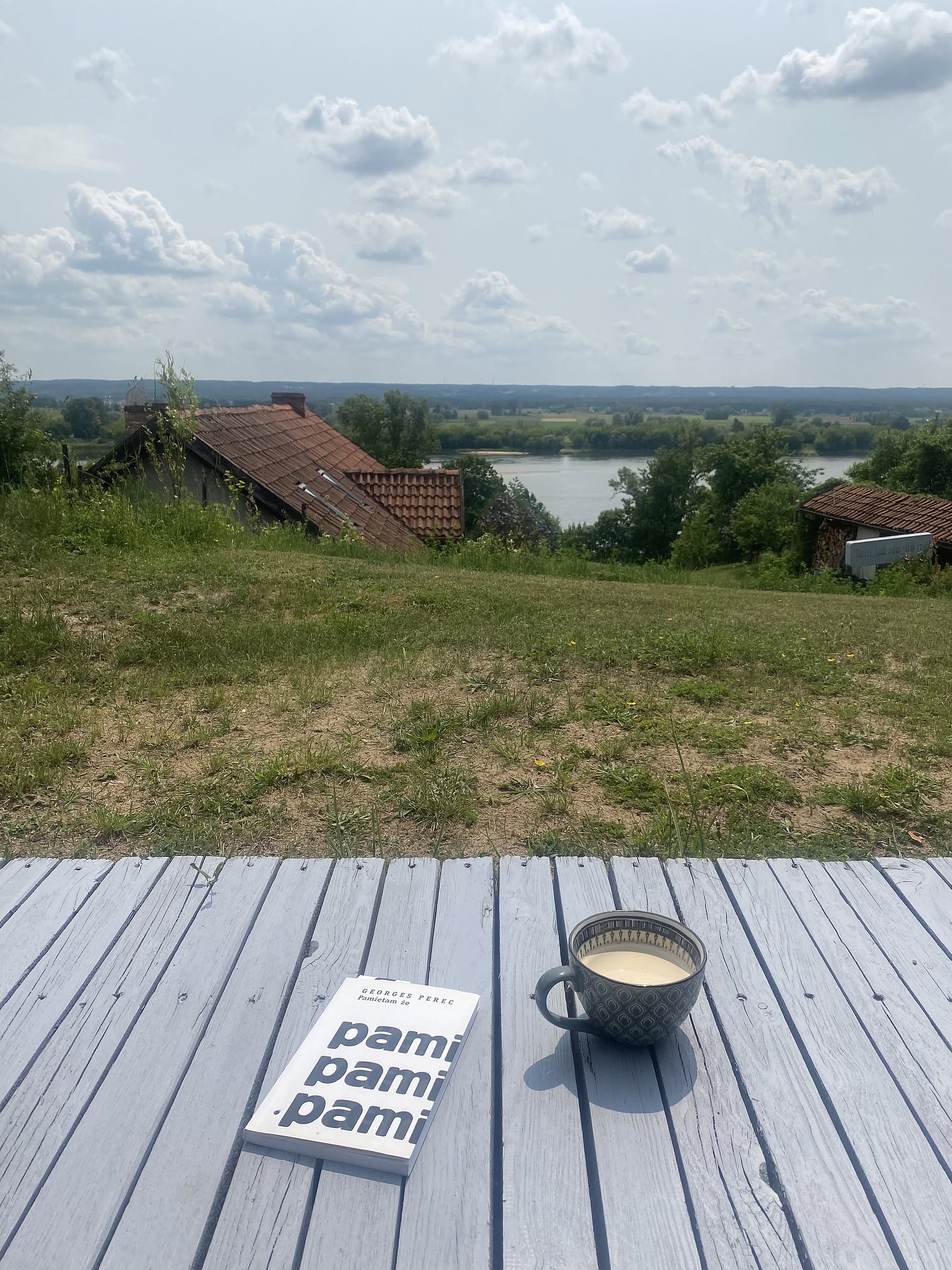 Drewniany taras, książka Pamietam, kawa, dom i rzeka w tle