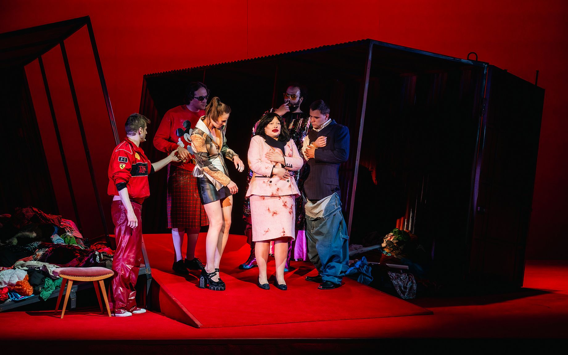 scena w teatrze, w środku kobieta w garsonce z przesadnym makijażem, obok gruba aktorów ubrana w dziwe stroje, czerwone światło