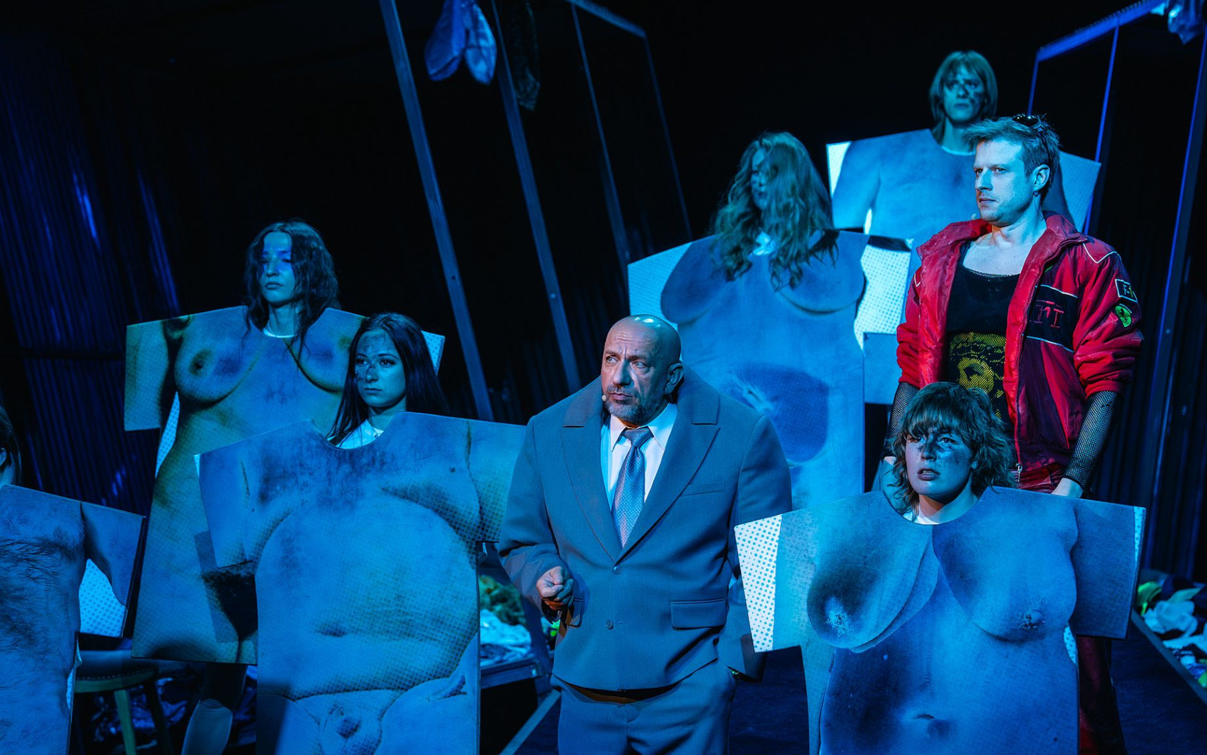 scena w teatrze, mężczyzna w czerwonym dresie, obok postaci w kostiumach udających ciało, niebieskie światło