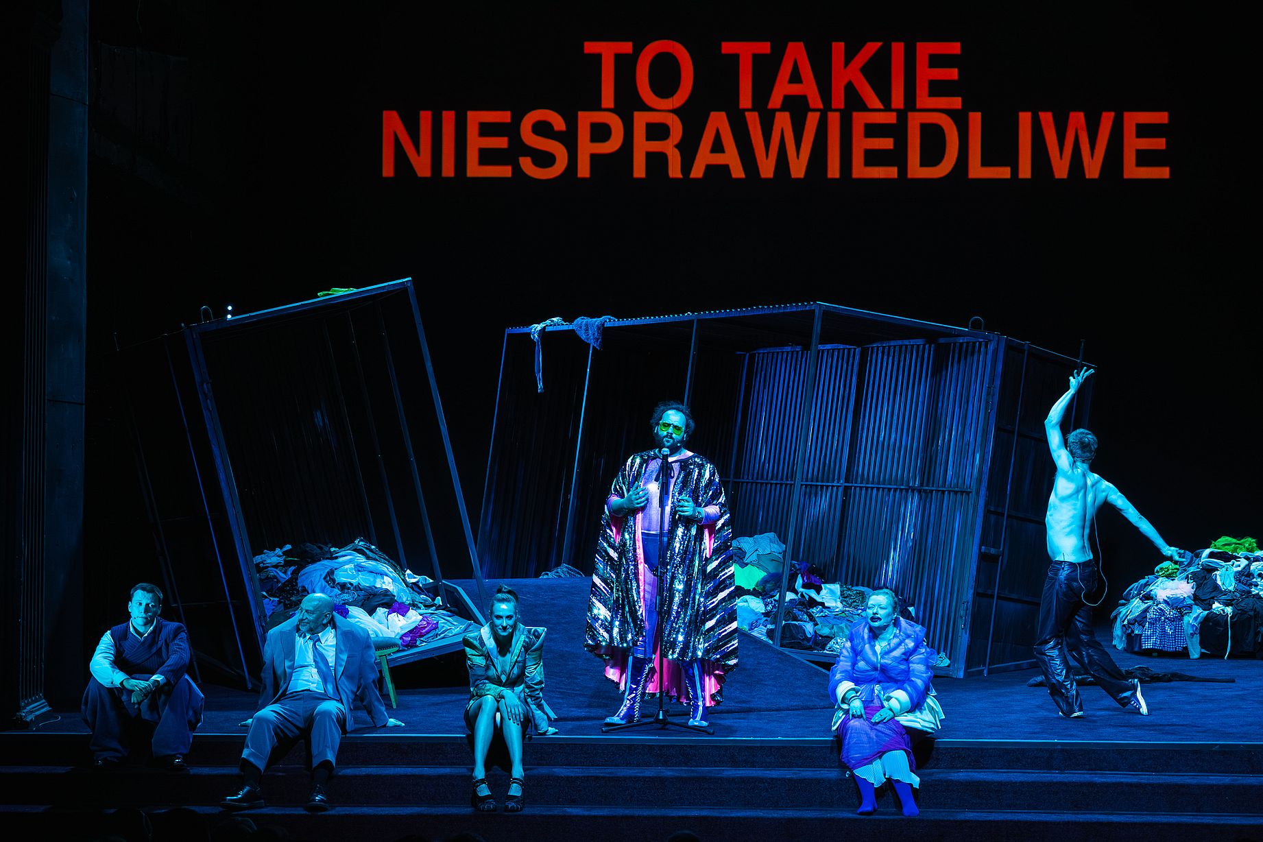 scena w teatrze, w środku mężczyzna w dziwnym kostimie, obok kilka postaci siedzi, jeden z mężczyzn bez koszuli tańczy, niebieskie światło