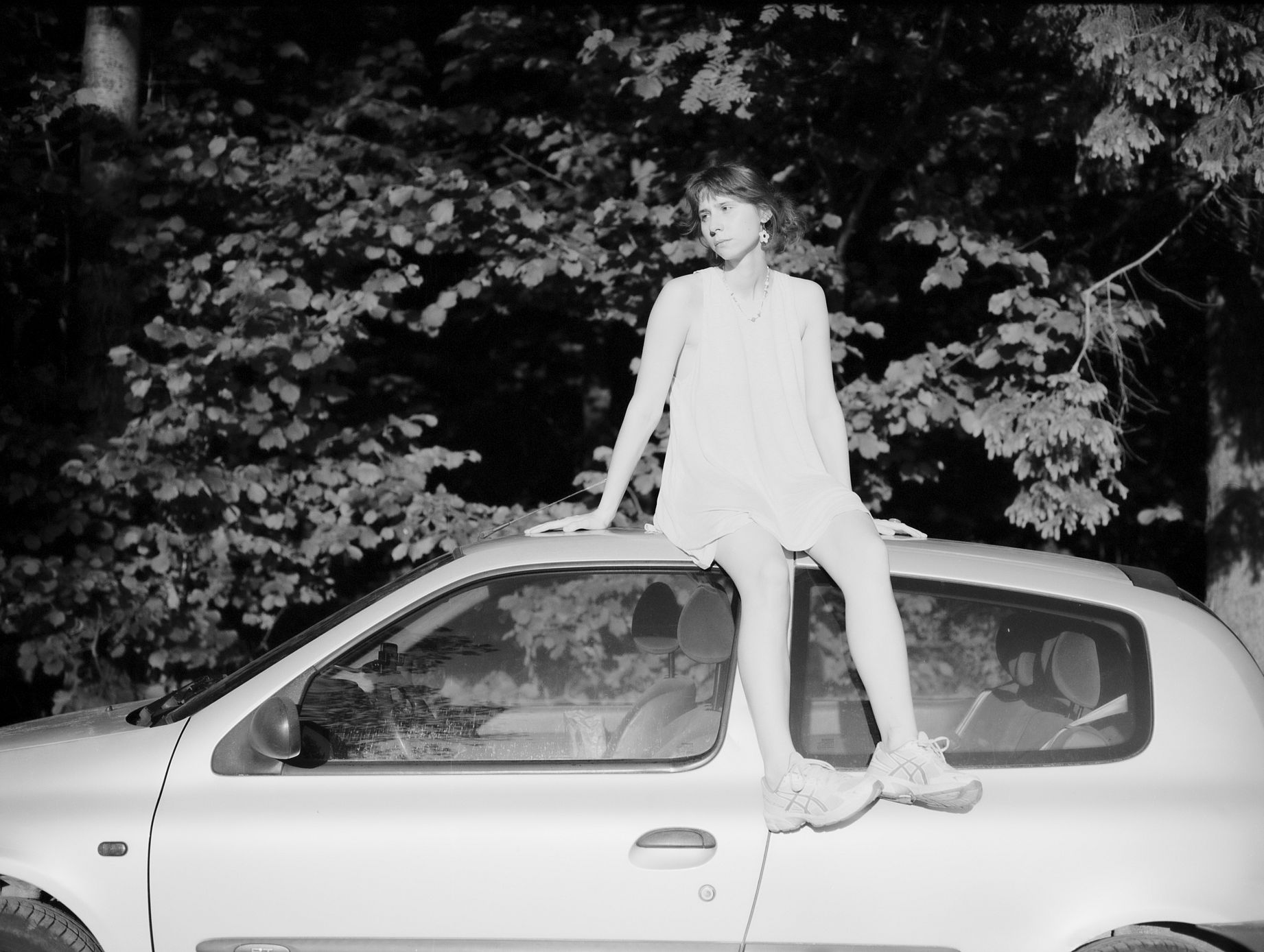 Czarno-białe zdjęcie plenerowe przedstawia kobietę siedzącą na dachu srebrnego samochodu, ubraną w jasną sukienkę i buty sportowe, na tle gęstych zar