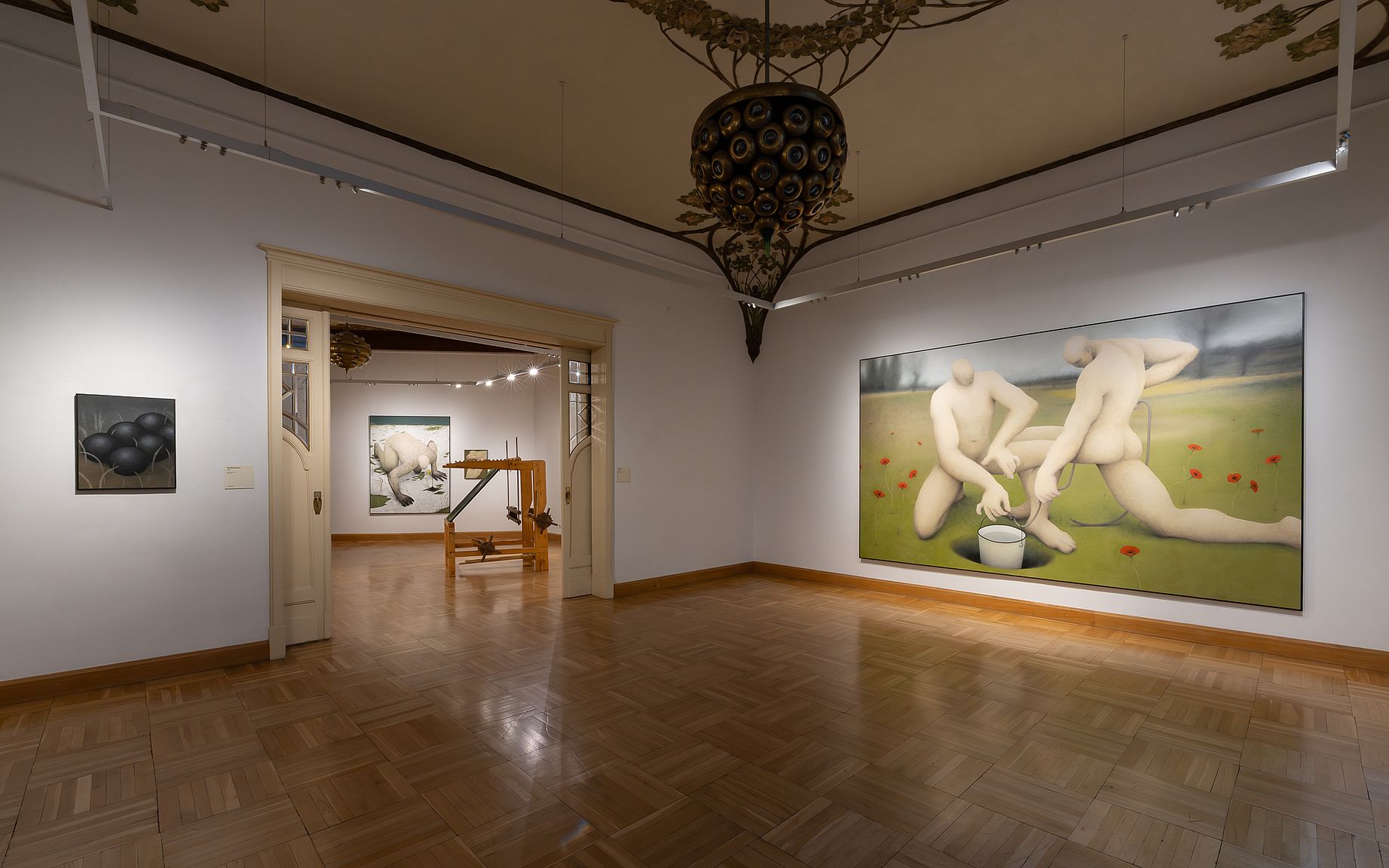 Galeria sztuki ma drewniany parkiet, białe ściany ze sztukaterią i kryształowy żyrandol, a na jednej ze ścian wisi duży obraz przedstawiający dwie nagie postacie klęczące i trzymające wiadro w trawie z czerwonymi kwiatami.