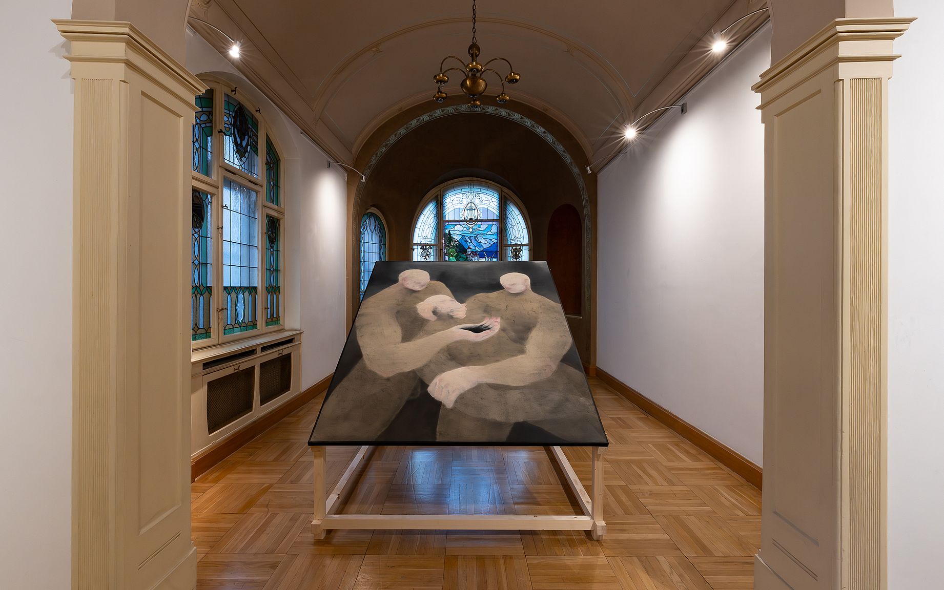 Obraz na drewnianym stelażu w galerii sztuki przedstawia dwie stylizowane, siedzące postaci w intymnej pozie, oświetlone przez górne światło i otoczone architektonicznymi detalami, takimi jak okna witrażowe i drewniane podłogi.