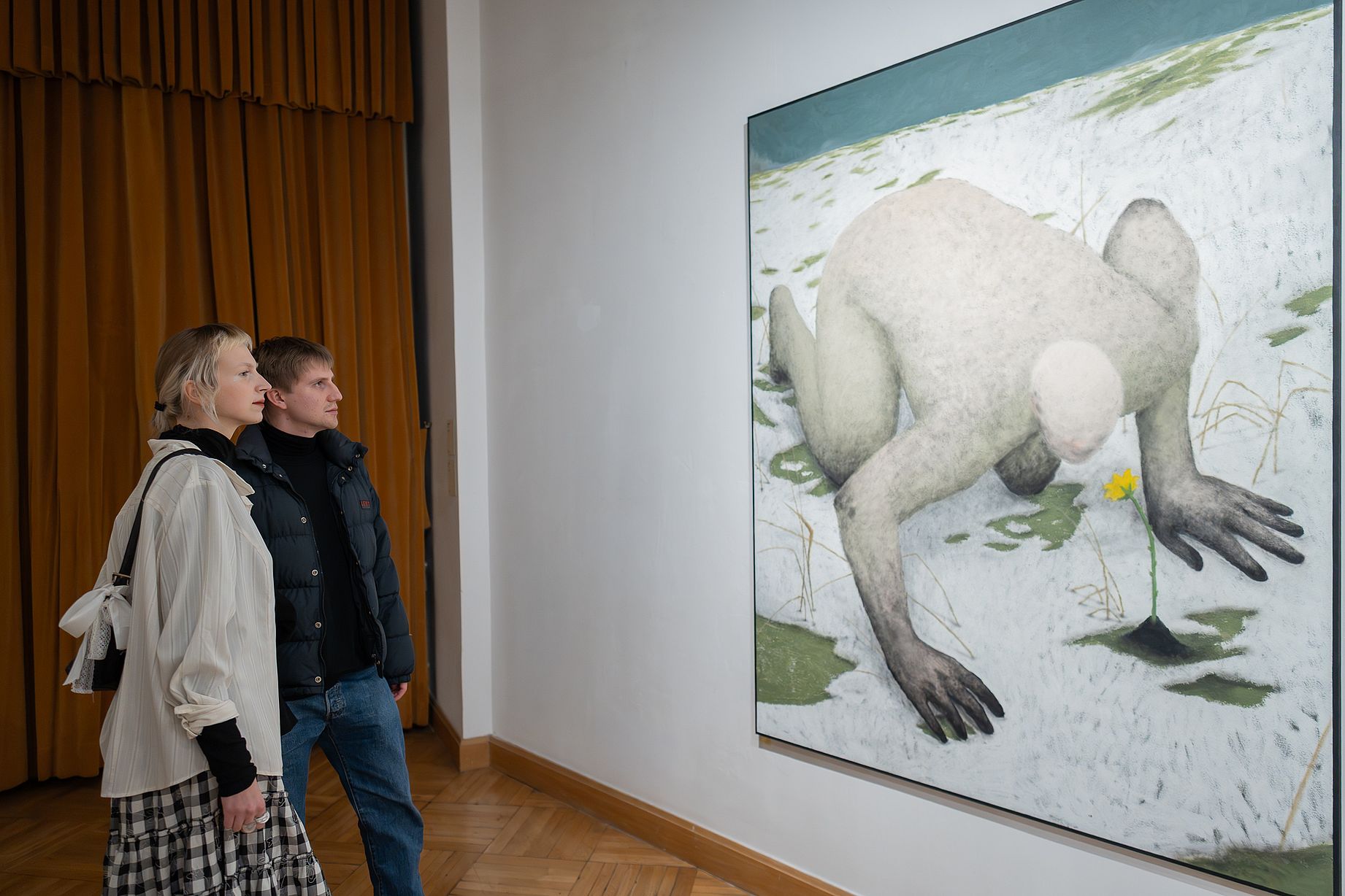 Kobieta i mężczyzna stoją w galerii sztuki, patrząc na duży obraz przedstawiający kucającą, jasną postać w zaśnieżonym krajobrazie z żółtym kwiatem.
