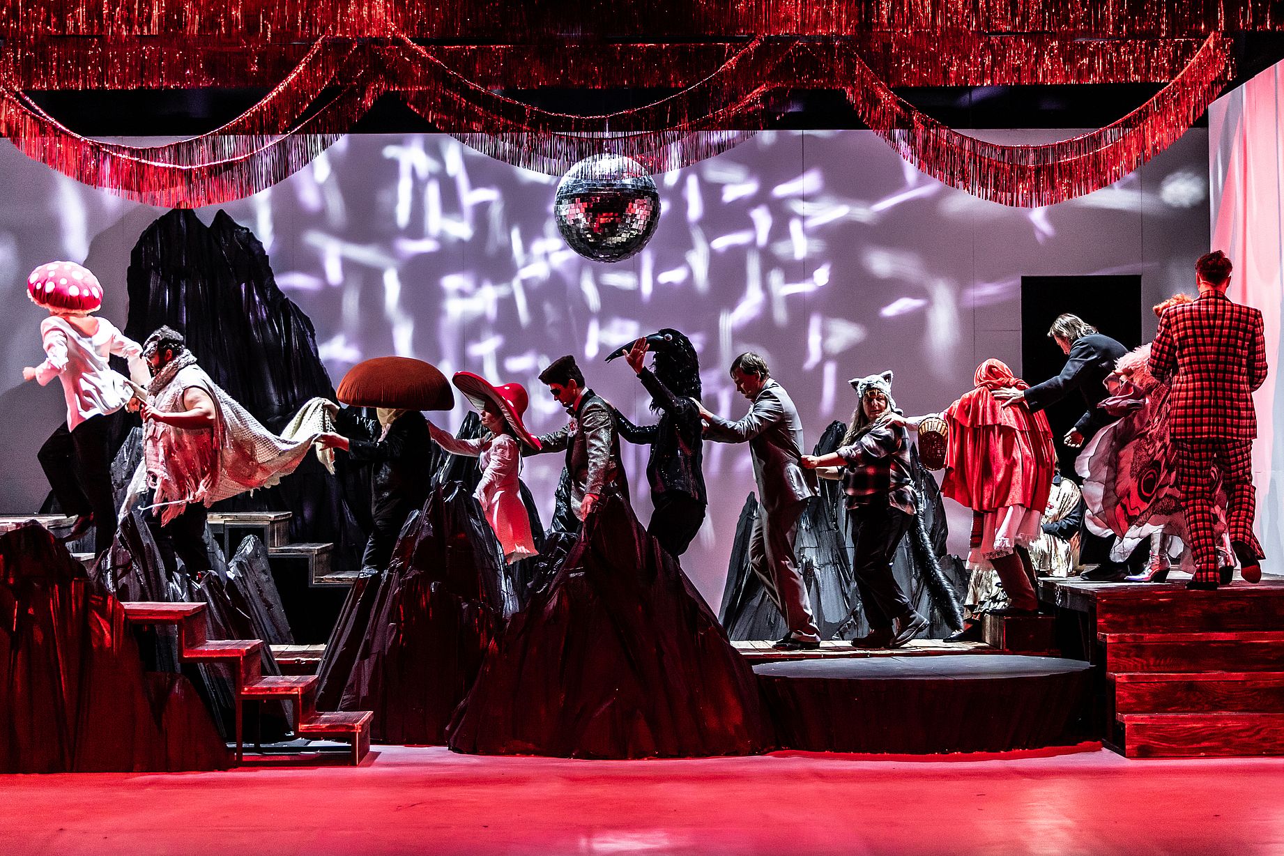 Scena teatralne, czerwone światło, dużo osób na scenie, czrne stroje, dziwny klimat, niektórzy aktorzy przebrani za zwierzęta