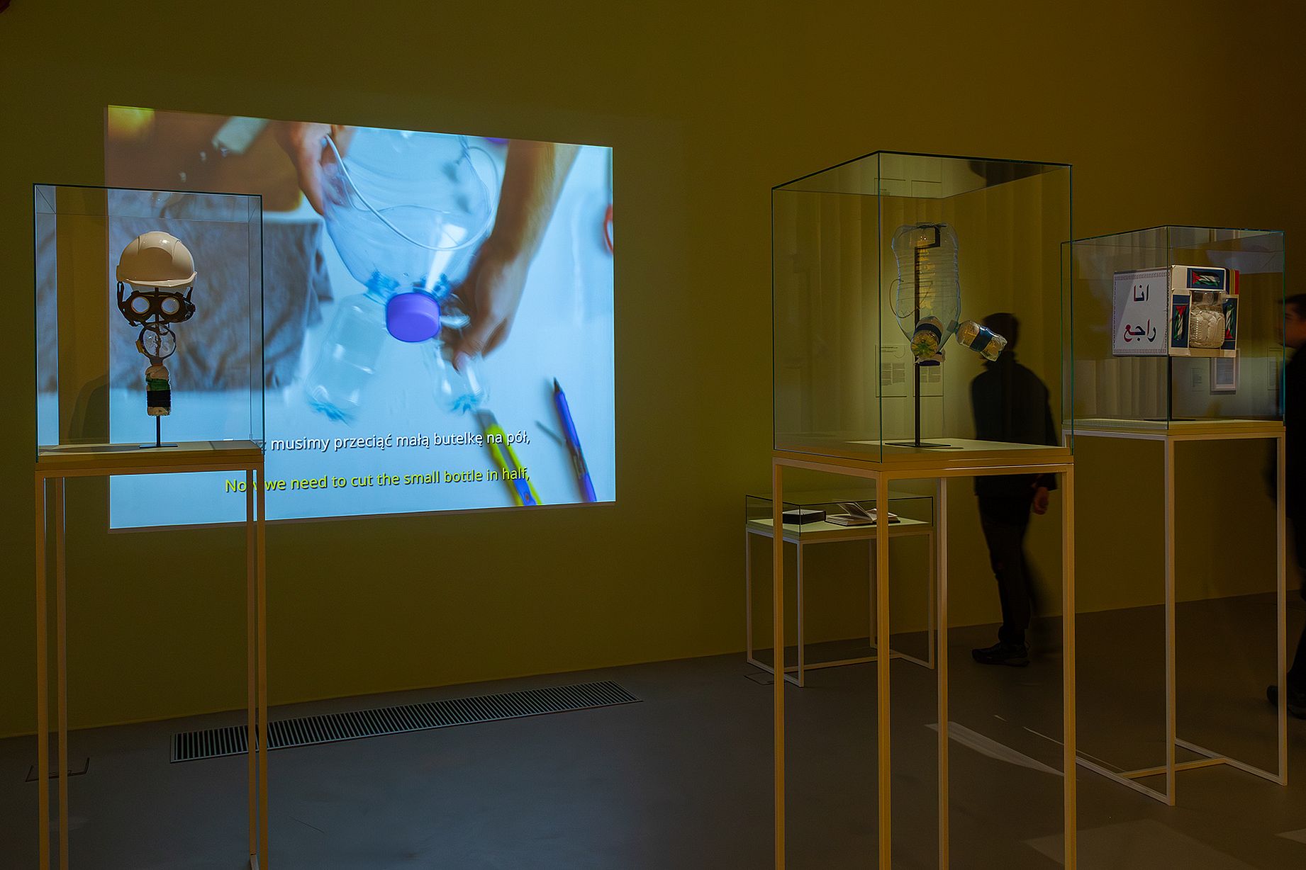 widok wystawy, projekcja, szklane gabloty,  żólte wysokie stoliki, widz w sali wystawowej 