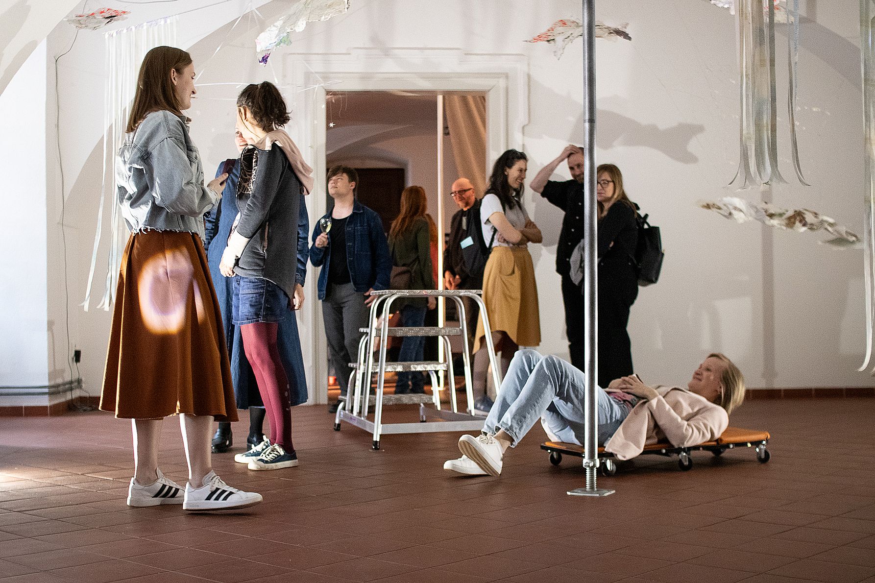 Na zdjęciu widać grupę osób oglądających wystawę sztuki współczesnej w jasnej sali, gdzie jedna osoba leży uśmiechnięta na niskim wózku pod instalacją wiszącą z sufitu, a inni stoją i rozmawiają wokół.