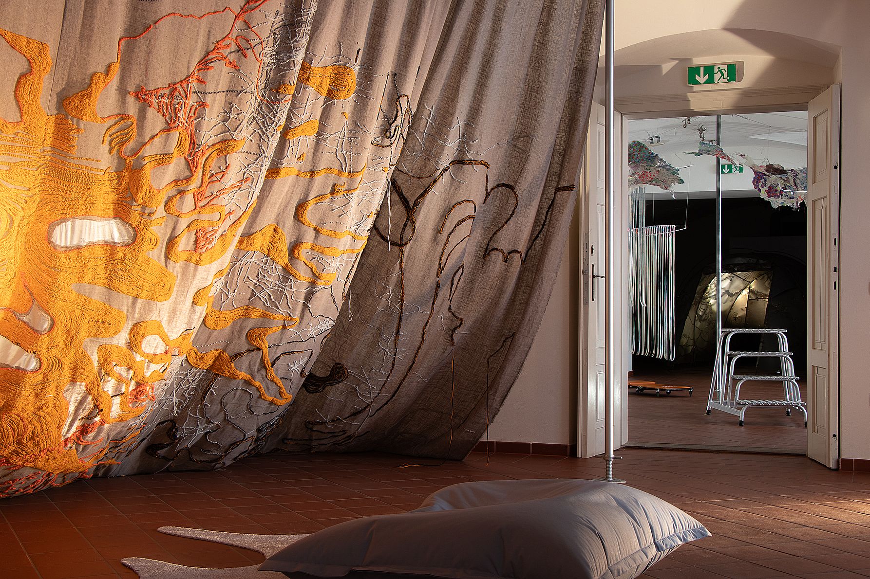 Na zdjęciu widać fragment sali wystawowej z dużą tkaniną w odcieniach pomarańczu i szarości zawieszoną na ścianie oraz instalacjami artystycznymi rozciągającymi się w kolejnych pomieszczeniach.