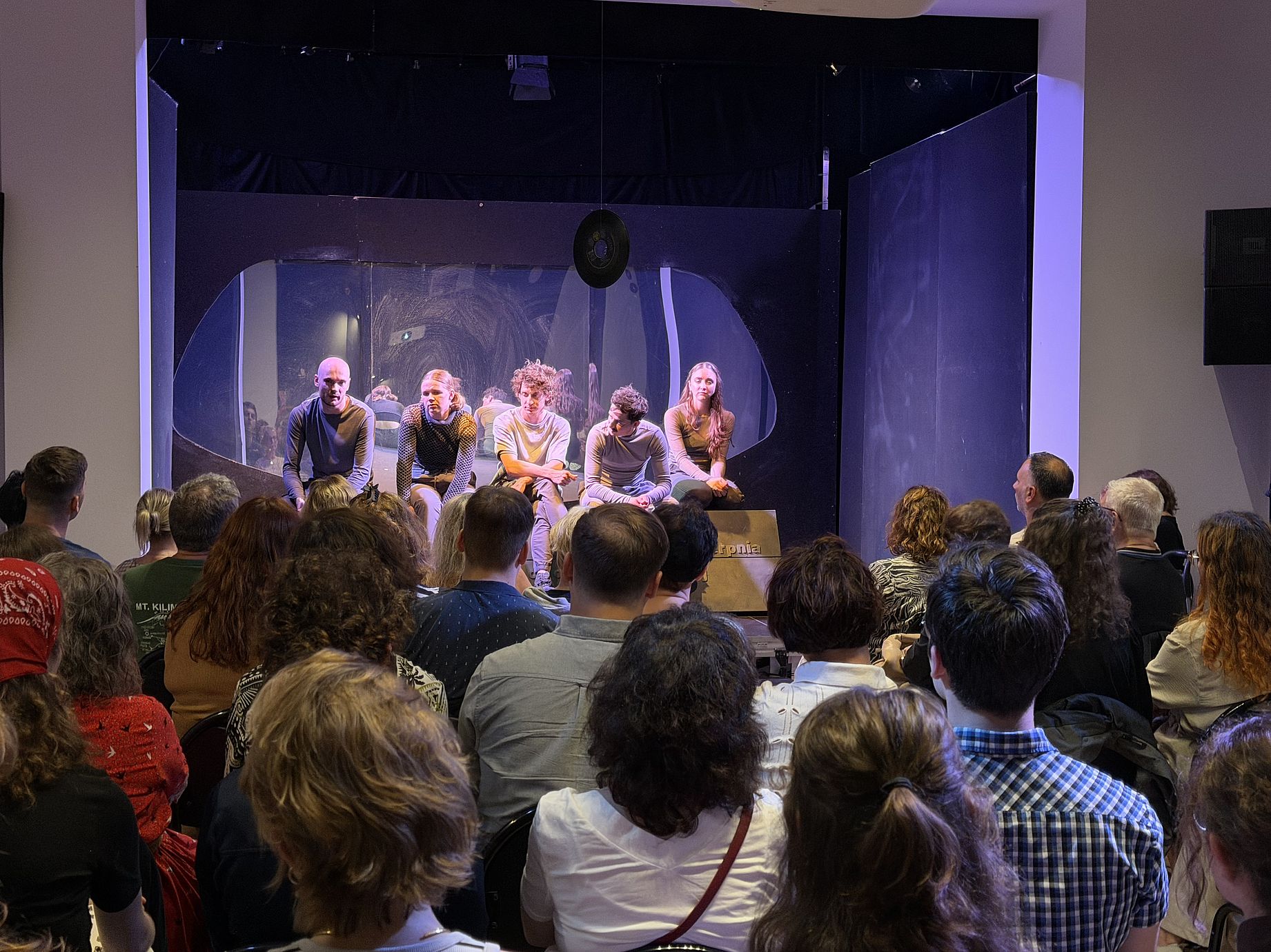 spektakl teatralnh, aktorzy siedzą na scenie, publiczność widziana z tyłu, fioletowe światło