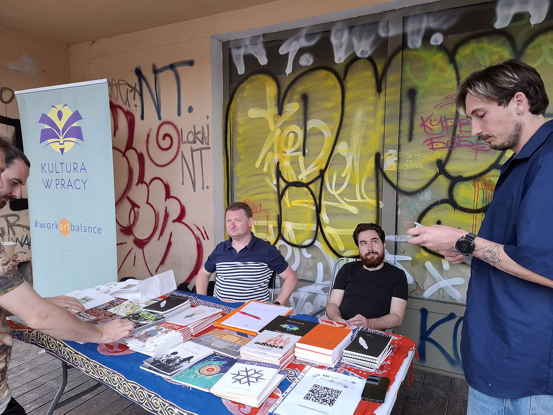 Pisanie wierszy na zamówienie na Placu Targowym, dwóch facetów siedzi przy stole pełnem książek, jeden stoi, patrzy w telefon