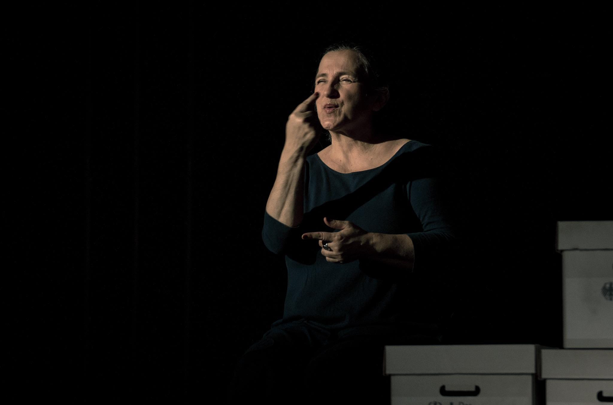 Kobieta siedzi w słabym świetle na tle ciemnej sceny i wykonuje gesty przypominające język migowy, mając obok siebie kilka kartonowych pudeł.