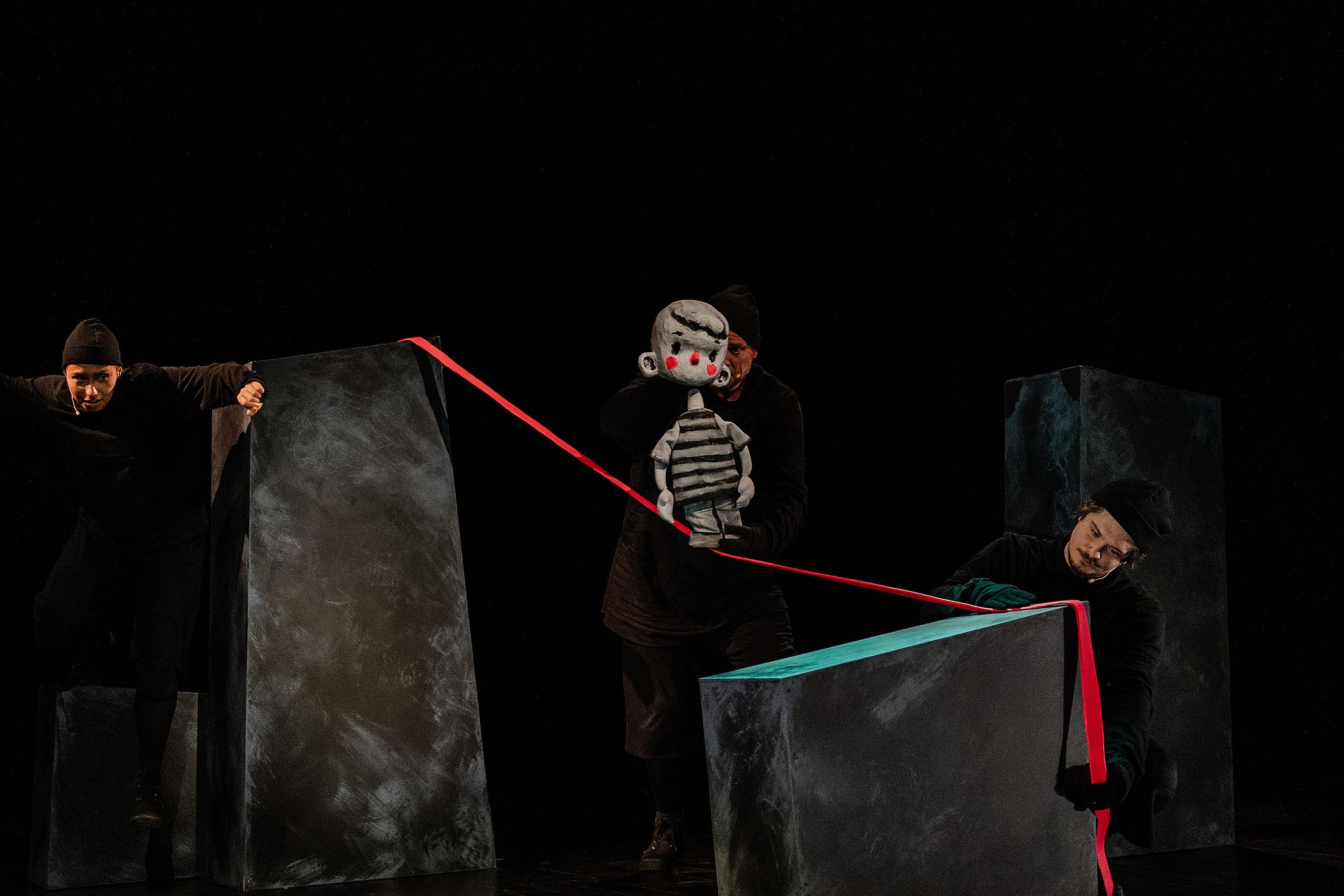 Na scenie w mrocznym oświetleniu trzech ubranych na czarno aktorów animuje dużą lalkę chłopca o szarym ciele i czerwonych policzkach, używając czerwonej wstążki rozciągniętej między ciemnymi blokami scenografii.