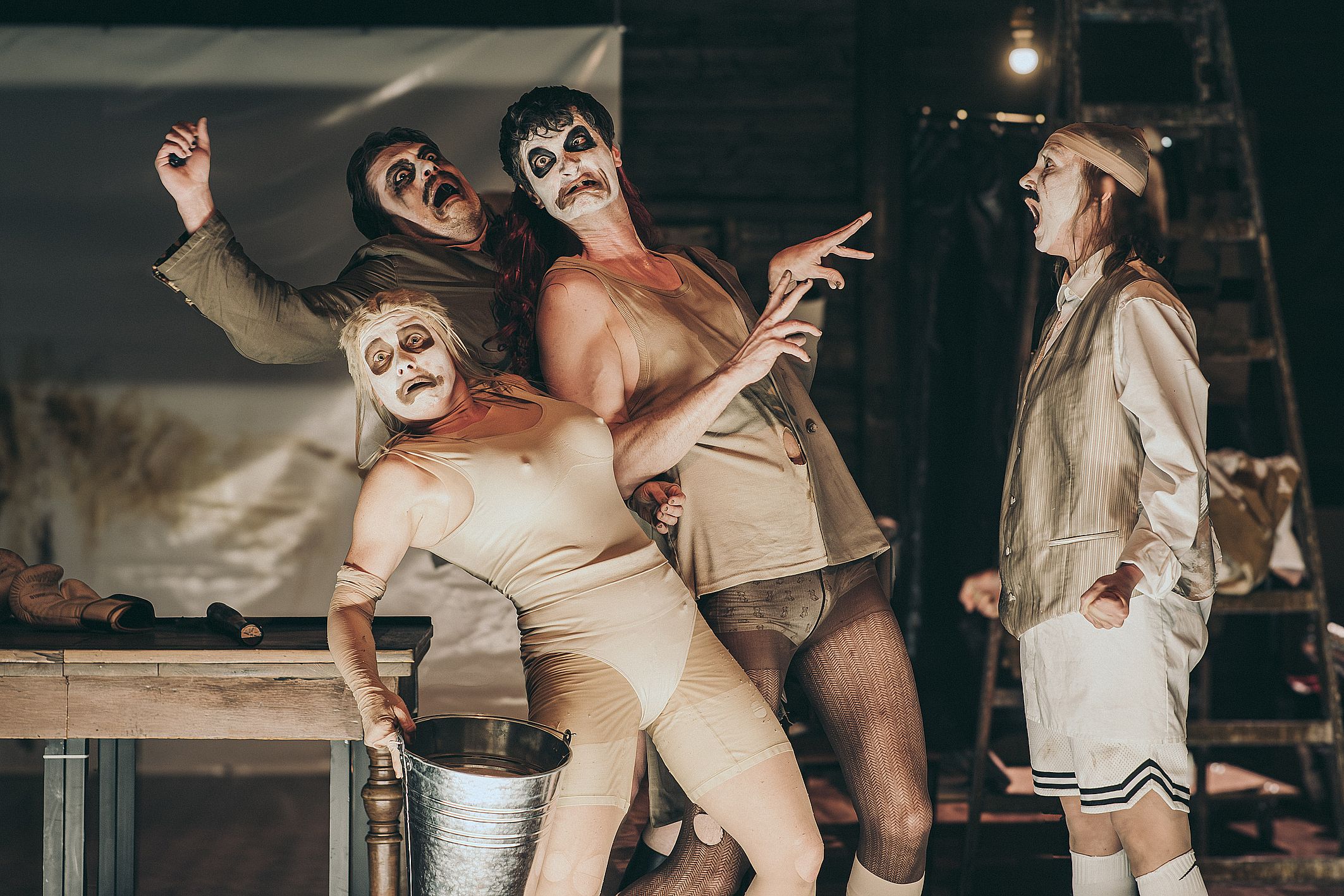 Czterech aktorów w groteskowym makijażu i dziwacznych kostiumach, zastygłych w przerysowanych pozach, tworzy na scenie ekspresyjną, pełną napięcia i komizmu scenę teatralną.