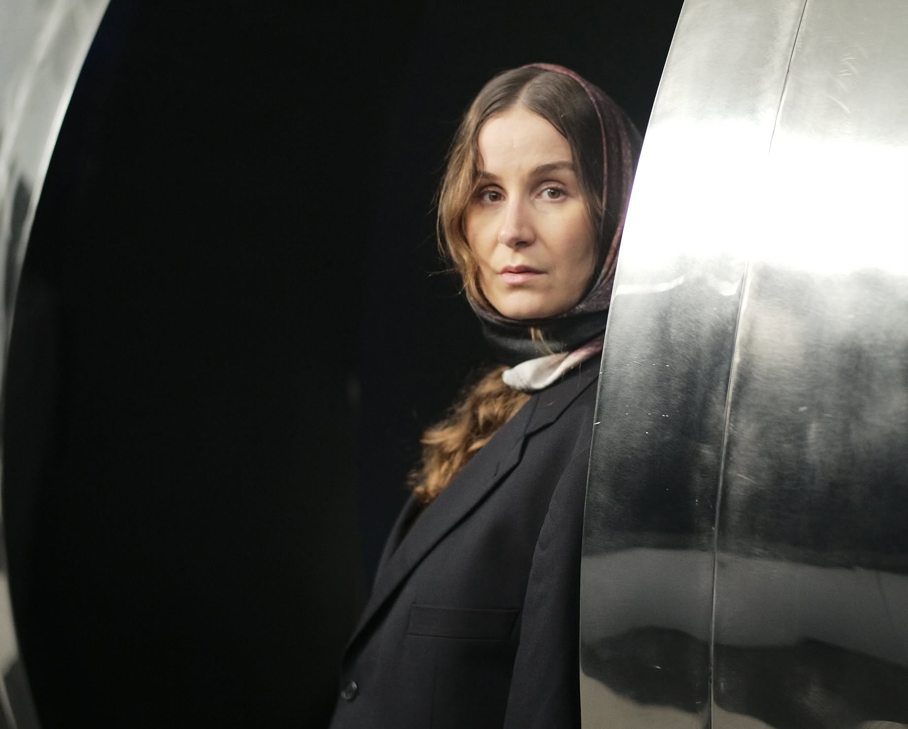 Kobieta w chustce na głowie, ubrana na czarno, stoi w środku instalacji artystycznej