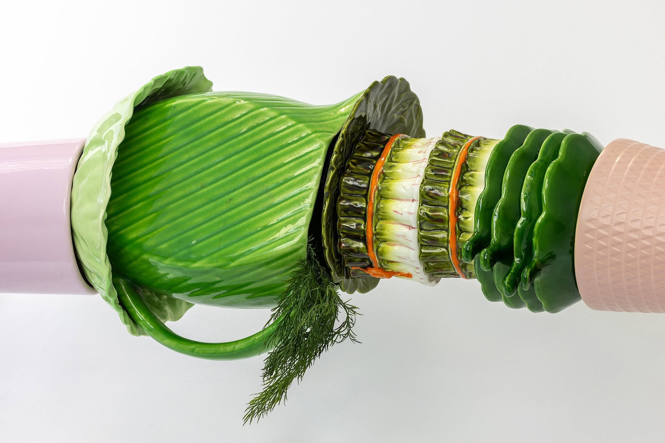 Obraz przedstawia surrealistyczną, poziomą rzeźbę ceramiczną, która łączy elementy różowej zastawy stołowej z realistycznie wymodelowanymi warzywami, takimi jak kapusta i koper, tworząc wrażenie hybrydowego, segmentowego obiektu.