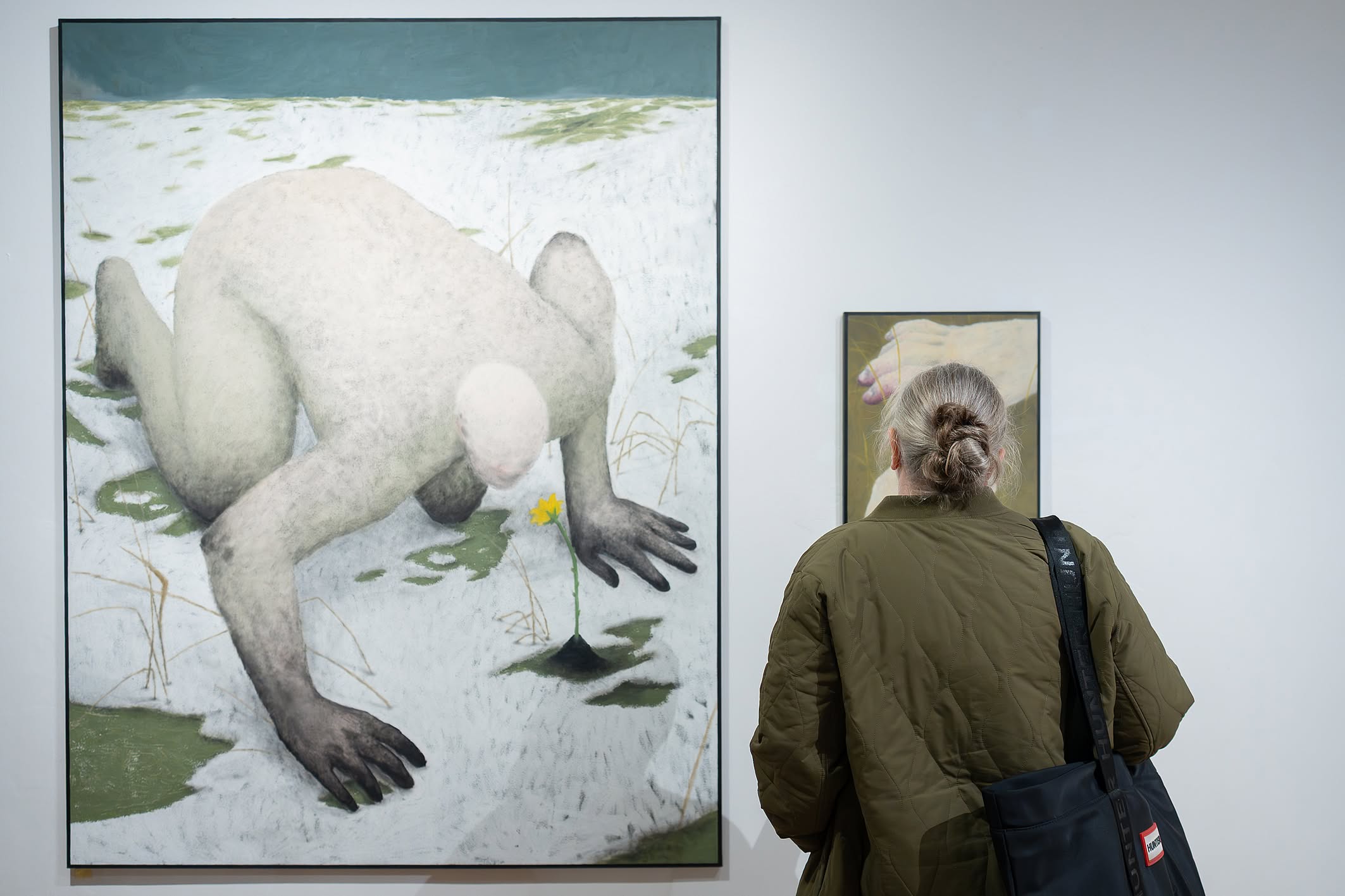  Kobieta stoi tyłem do kamery, patrząc na duży obraz przedstawiający kucającą postać w zaśnieżonym krajobrazie zbliżającą się do małego żółtego kwiatu.