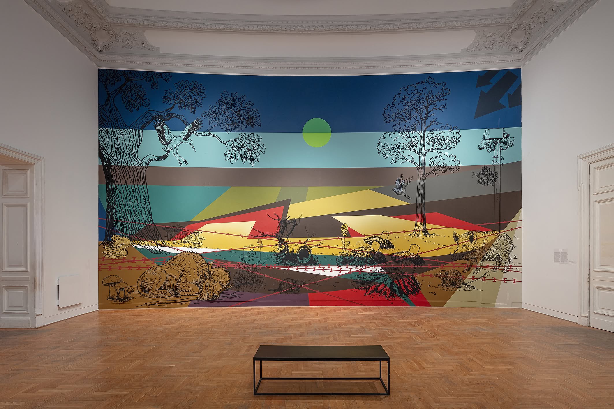 Wielobarwny mural autorstwa kolektywu Aravani Art Project przedstawia kilka kobiet w tradycyjnych strojach, otoczonych dużymi kwiatami i ptakiem, w pomieszczeniu zaaranżowanym z poduszkami i patchworkowymi dywanami na podłodze