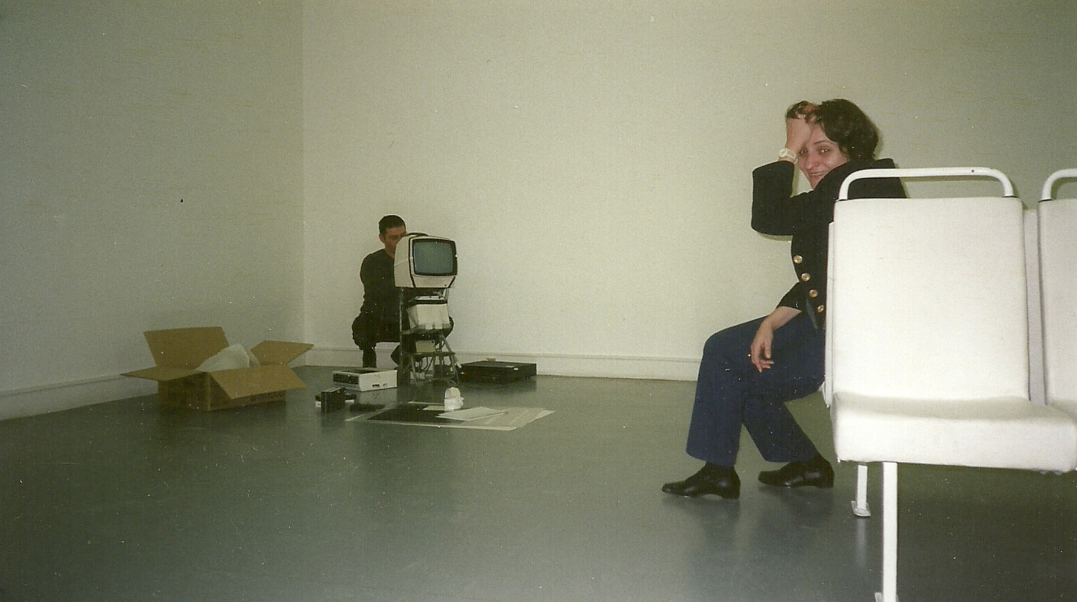 Na zdjęciu w pustym, jasnym pokoju siedzi kobieta uśmiechająca się do aparatu, a w tle mężczyzna klęczy przy starym monitorze i kartonie.