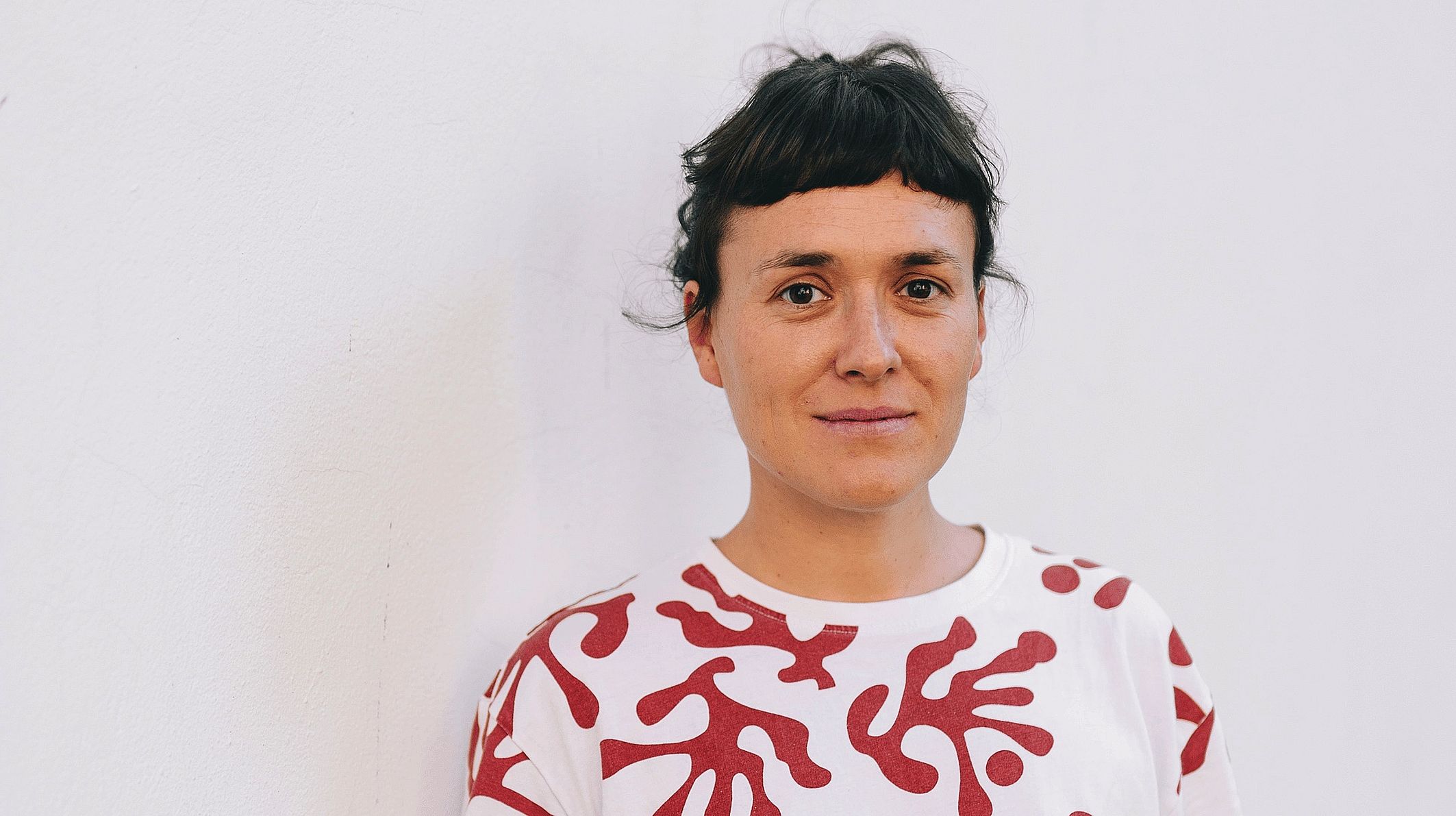 lekko uśmiechnięta kobieta patrzy obrana w białą blukę z czerwono-brązowe plamy w obiektyw, 