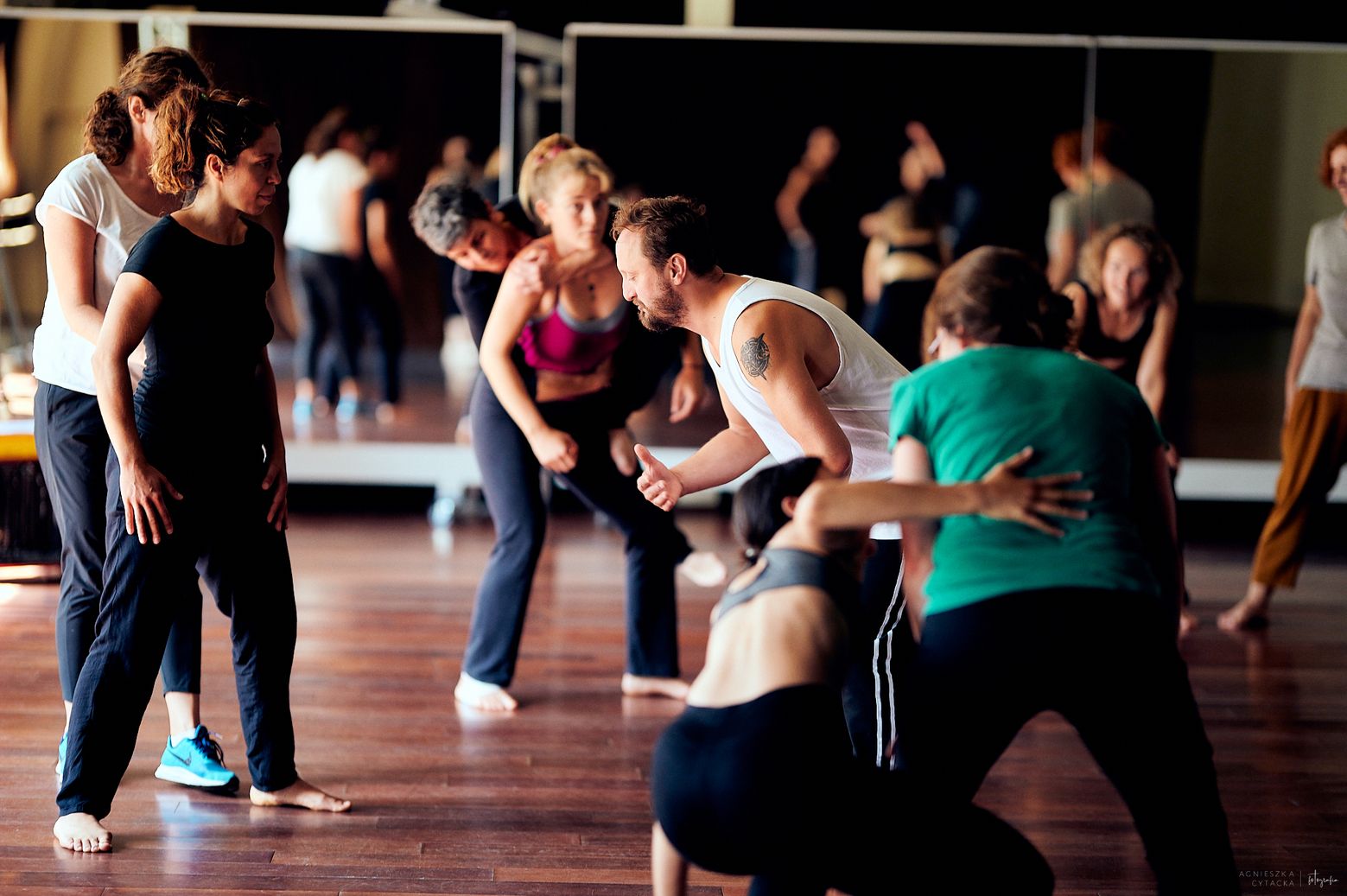 Na zdjęciu widać grupę osób uczestniczących w intensywnych warsztatach ruchowych lub tanecznych – są skupieni, w różnych pozach ciała, w trakcie wspólnego ćwiczenia, a centralną postacią jest mężczyzna w białej koszulce, który energicznie instruuje pozost