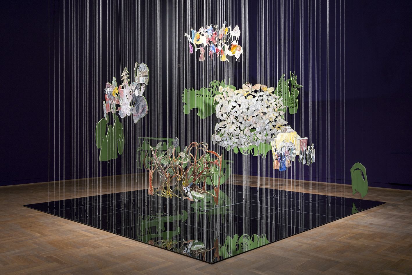 Na zdjęciu widać dużą instalację artystyczną w pomieszczeniu oświetlonym światłem studyjnym, na tle ciemnoniebieskiej ściany, z jasną, drewnianą podłogą i kwadratowym, czarnym, lustrzanym podestem, na którym odbija się całe dzieło. Instalacja składa się z kilkudziesięciu wyciętych, płaskich figur wykonanych z różnych materiałów, głównie zielonego, nieprzezroczystego tworzywa i białego, puszystego materiału przypominająceg