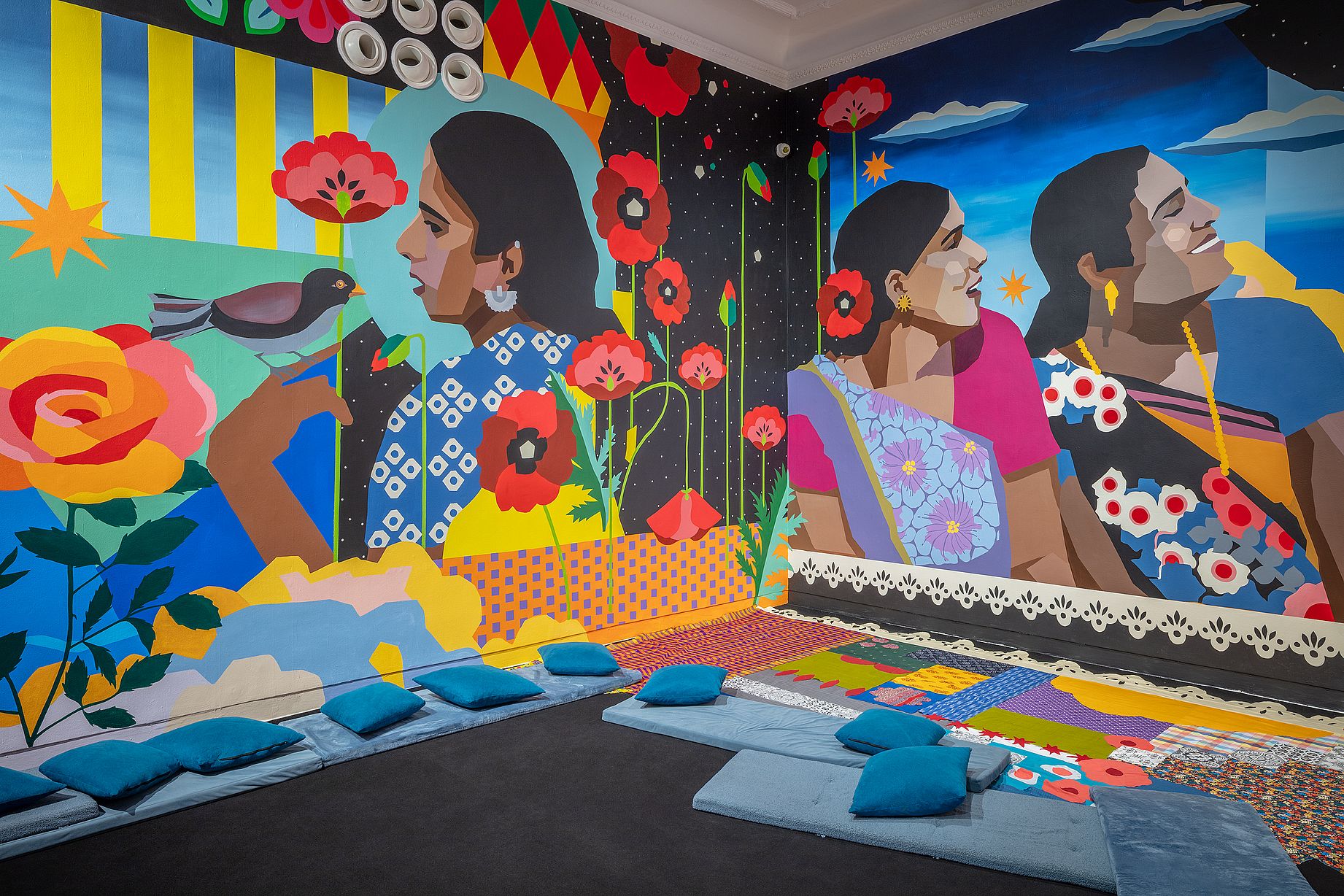 Wielobarwny mural autorstwa kolektywu Aravani Art Project przedstawia kilka kobiet w tradycyjnych strojach, otoczonych dużymi kwiatami i ptakiem, w pomieszczeniu zaaranżowanym z poduszkami i patchworkowymi dywanami na podłodze. 